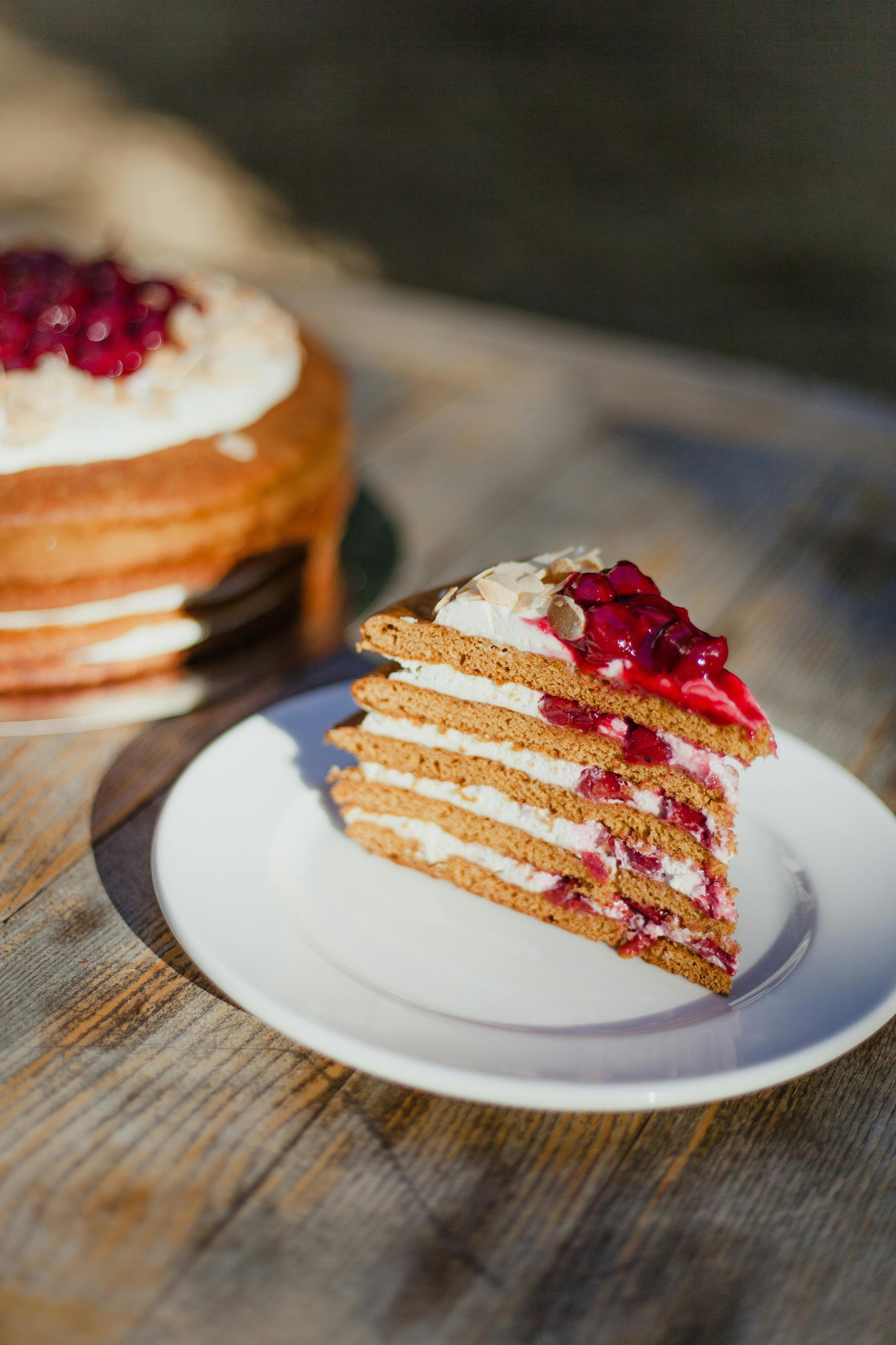 A slice of layered cake with cream and red fruit topping on a white plate, with the remaining cake in the background on a wooden surface.