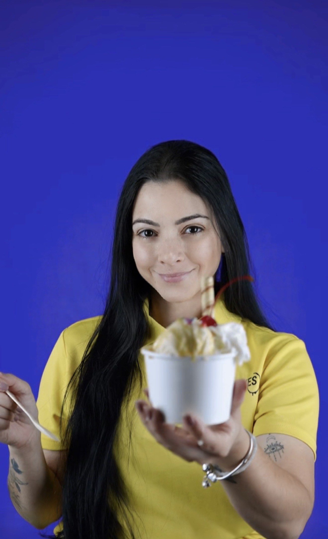 A woman in a yellow shirt holding a bowl of ice cream with whipped cream, caramel syrup, a cherry, and a wafer stick, smiling at the camera against a blue background.