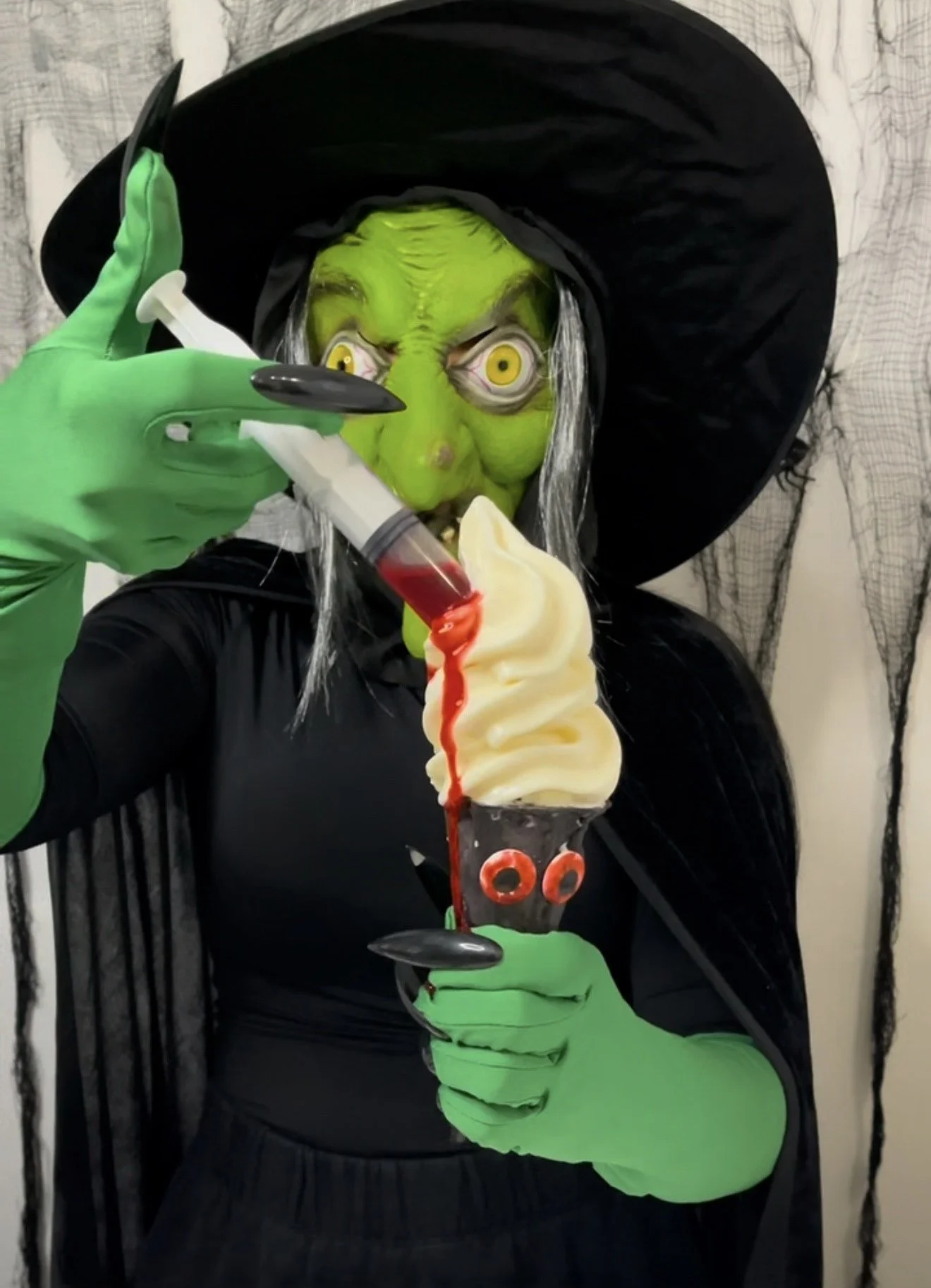 A person dressed as a witch with green face makeup, long gray hair, and a large black hat, holding an ice cream cone decorated with eyes, while applying a syringe filled with red liquid to the ice cream. It's a Party Bites MIA event catering.