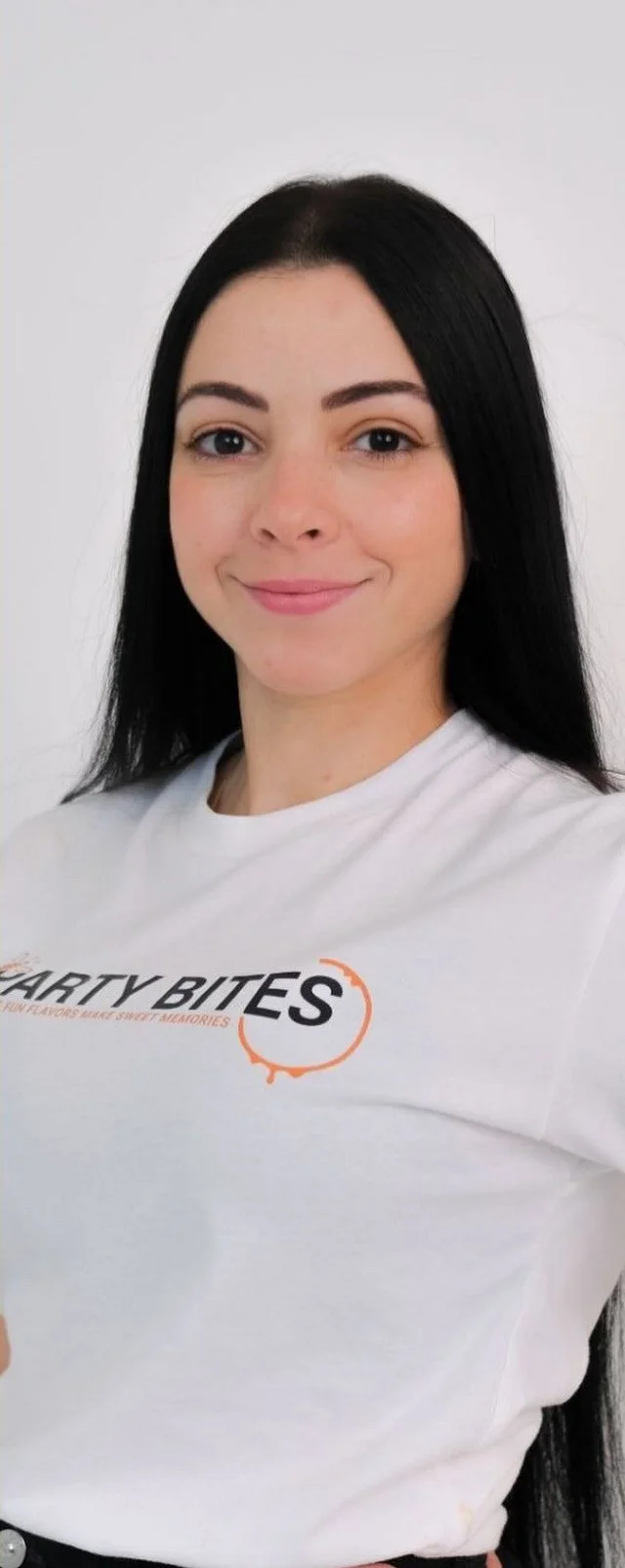 A woman with long black hair wearing a white t-shirt that reads 'Party Bites' and is smiling, standing against a plain white background.