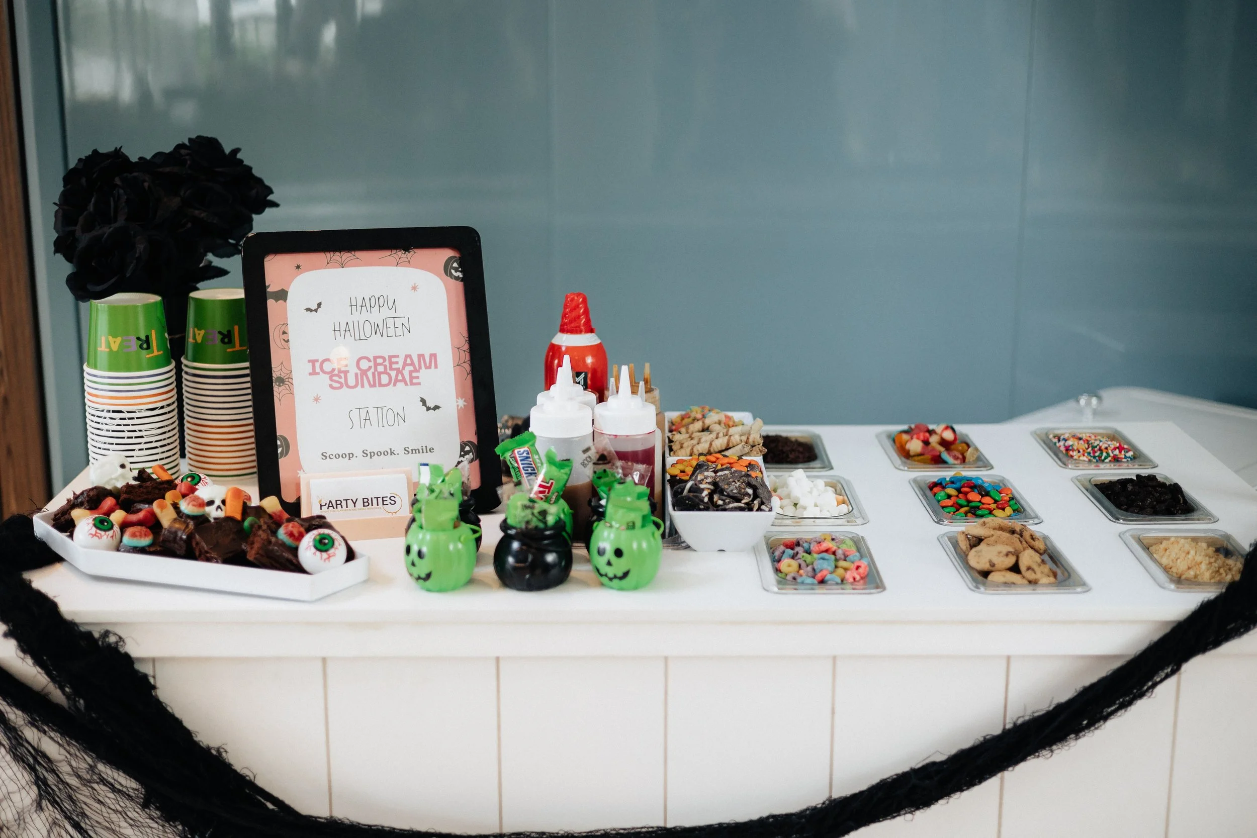 Halloween-themed ice cream sundae station with cups, party favors, and various toppings and candies.