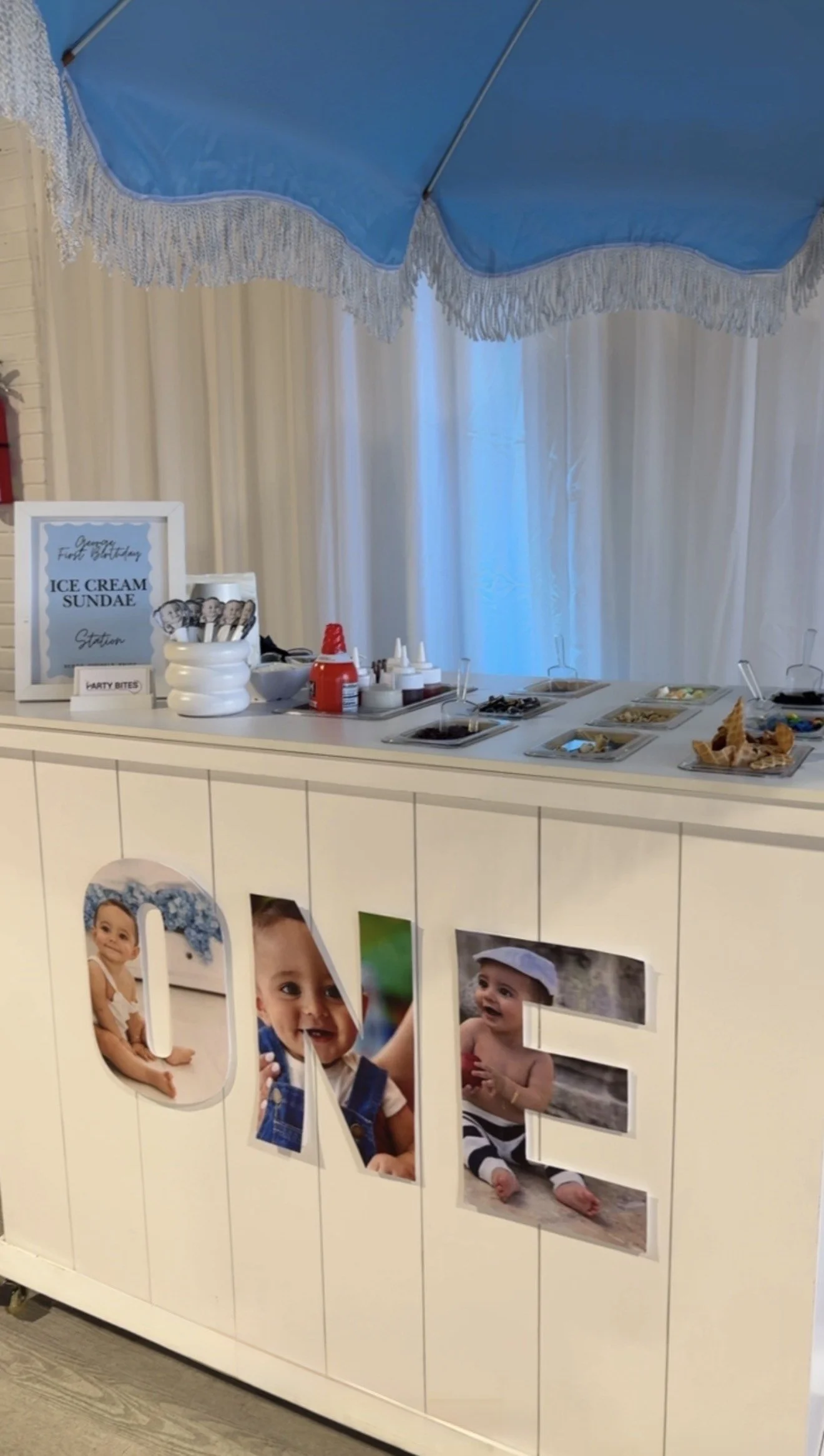 A birthday party dessert station with ice cream sundaes, toppings, and a sign that reads 'First Birthday'. The front of the station features the word 'ONE' decorated with photos of a baby. There is a large blue umbrella above the setup.