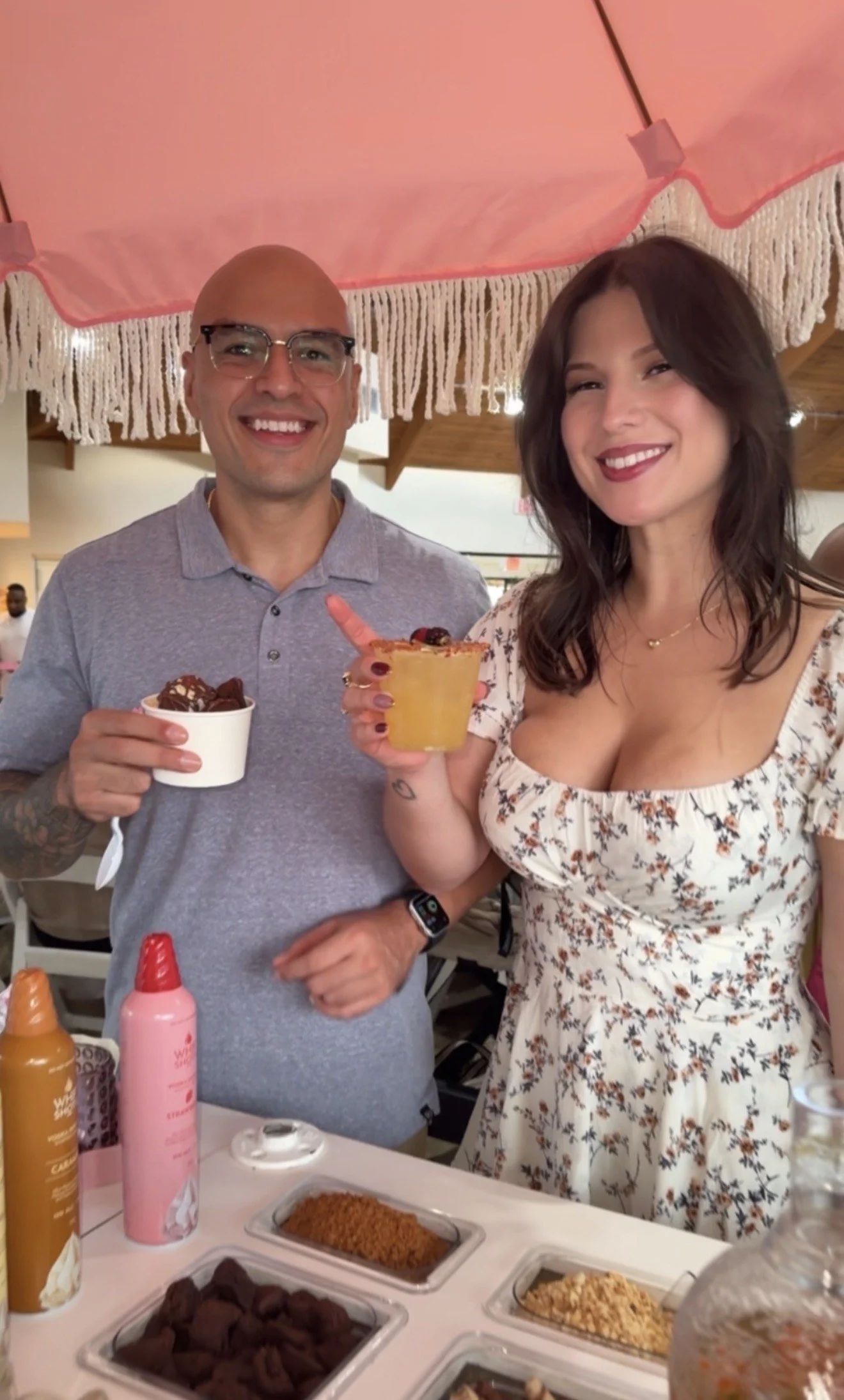A smiling man and woman standing together indoors, holding drinks and posing for a photo at a dessert or snack bar with various toppings and condiments on the table in front of them.