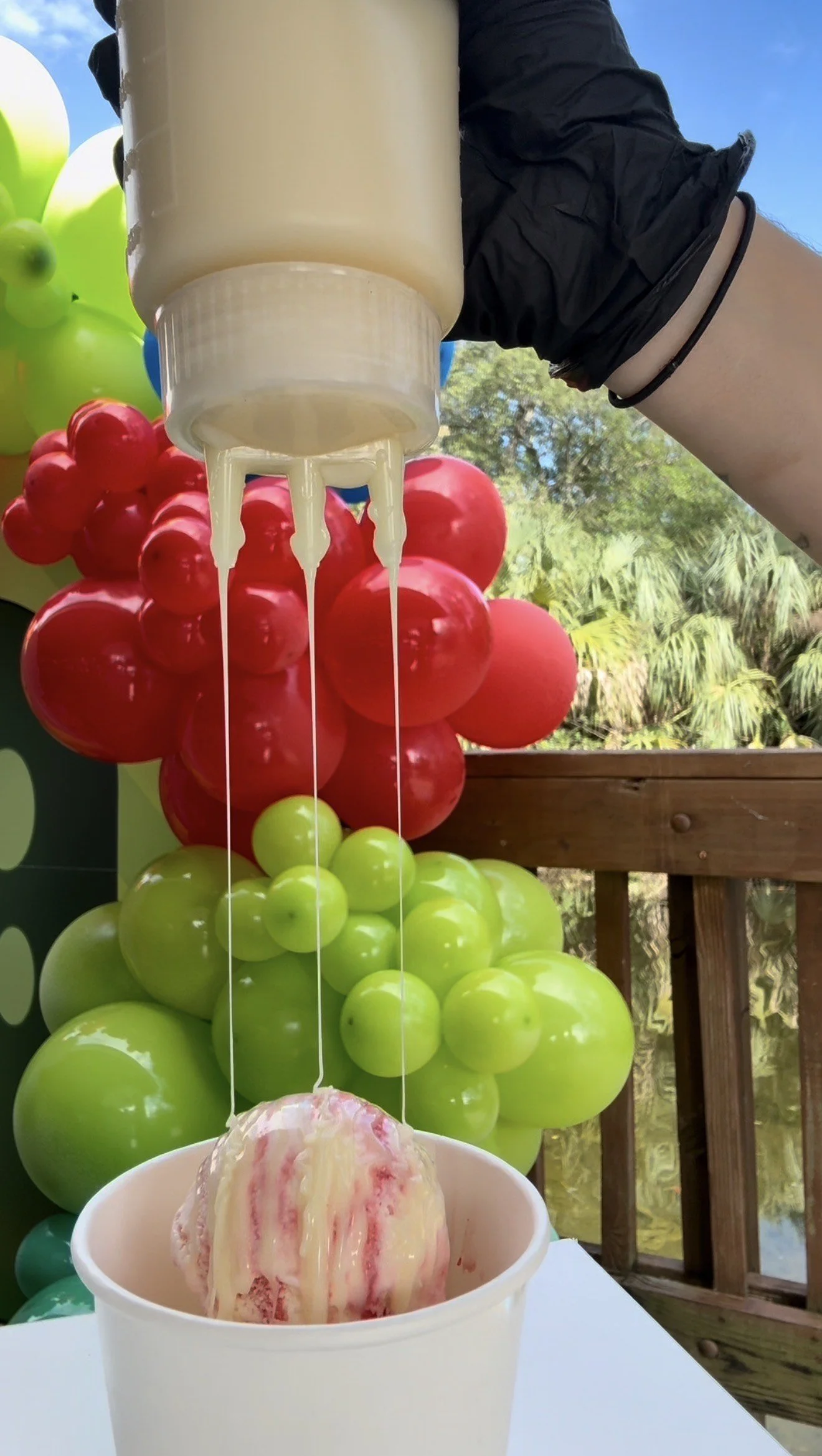 Ice cream with pink and white swirls in a paper cup, being drizzled with caramel sauce from a squeeze bottle, with colorful balloons in the background.
