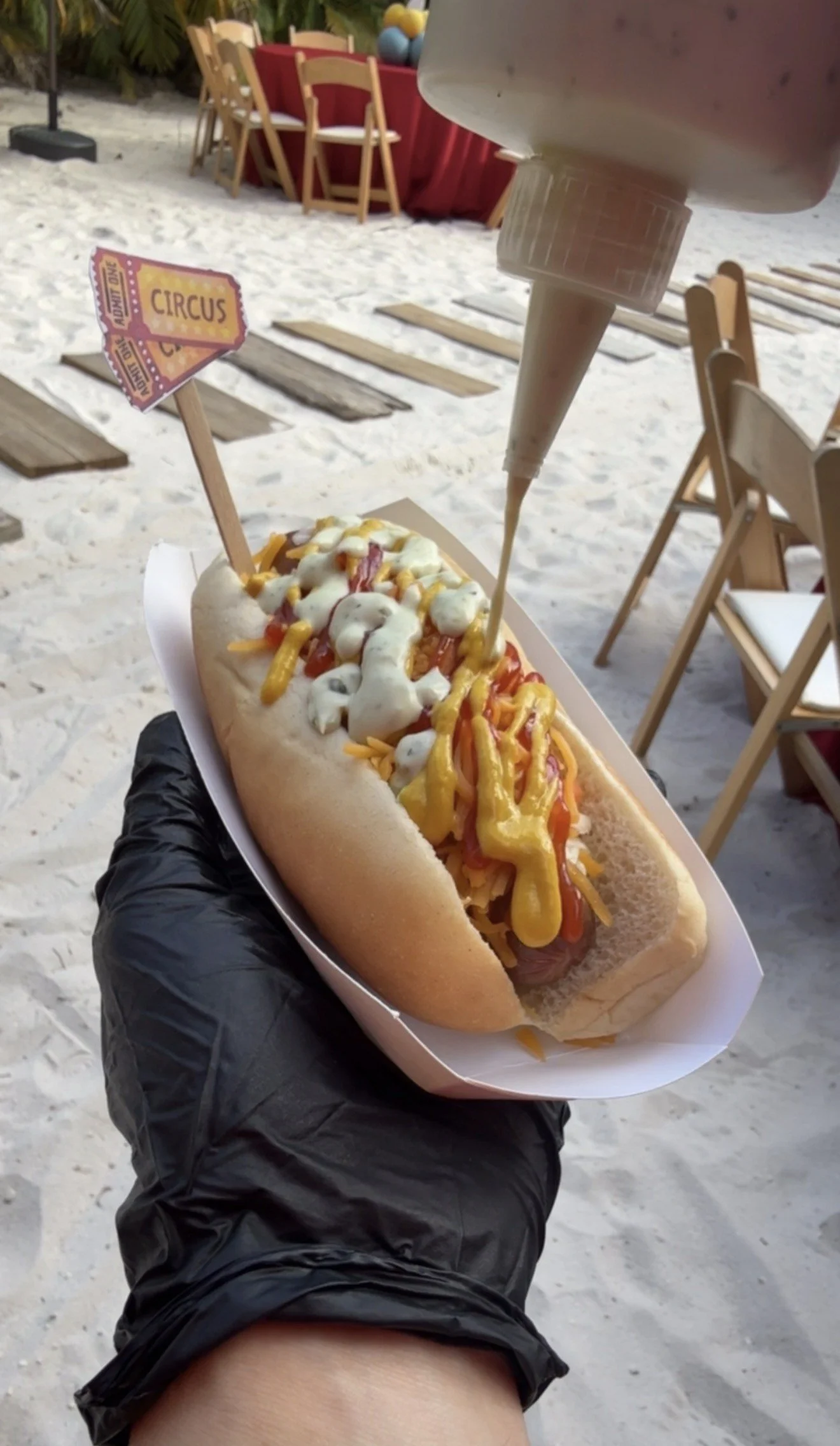 A hot dog topped with mustard, ketchup, and mayonnaise, served in a paper tray, with a squeeze bottle of mayonnaise above it, held by a person wearing a black glove at the beach, with tables, chairs, and a sign that reads 'Circus' in the background.
