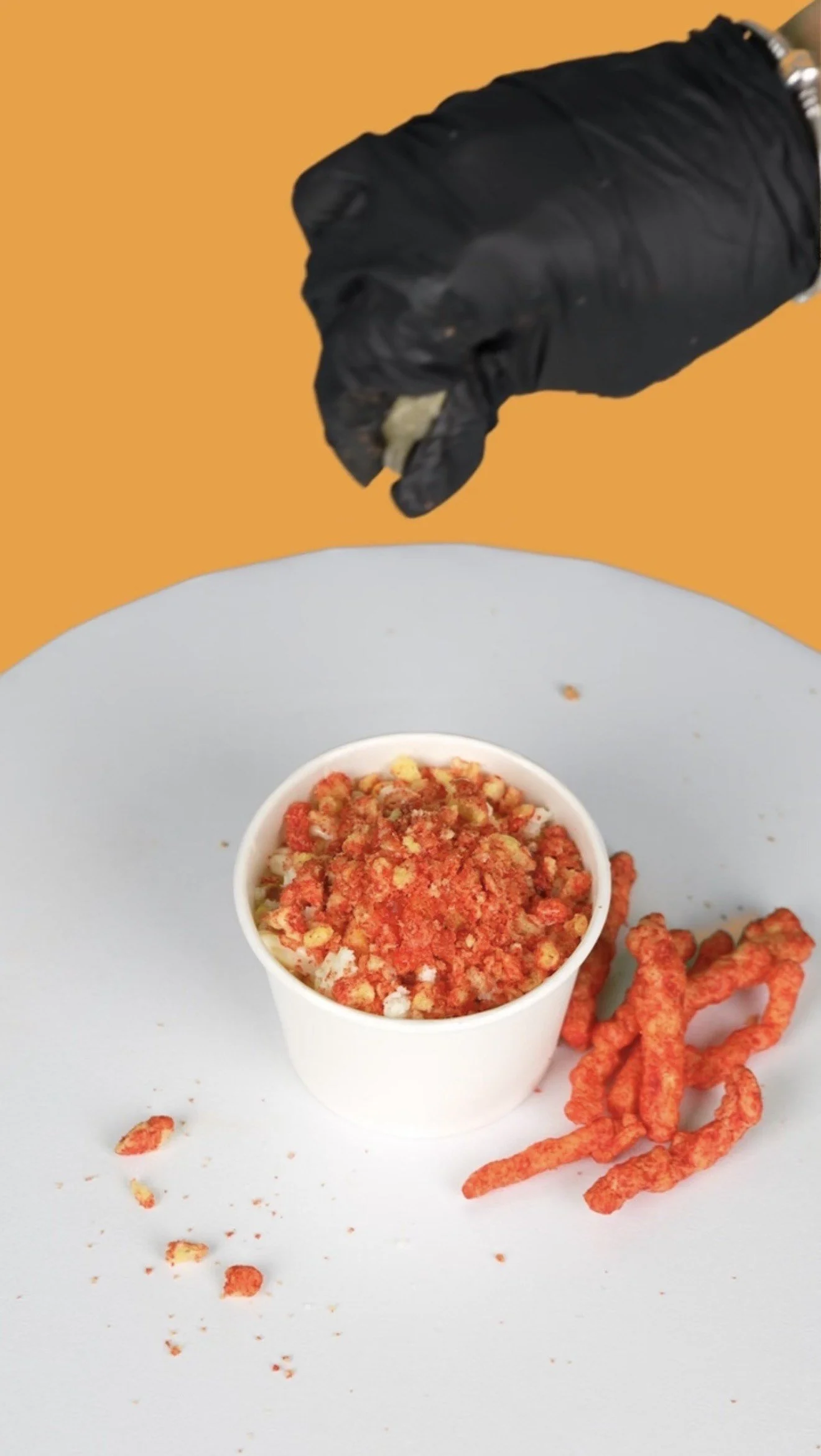 A hand wearing a black glove holds a snack over a white cup of crushed snack crumbs on a gray surface with orange background, some snacks are on the surface.