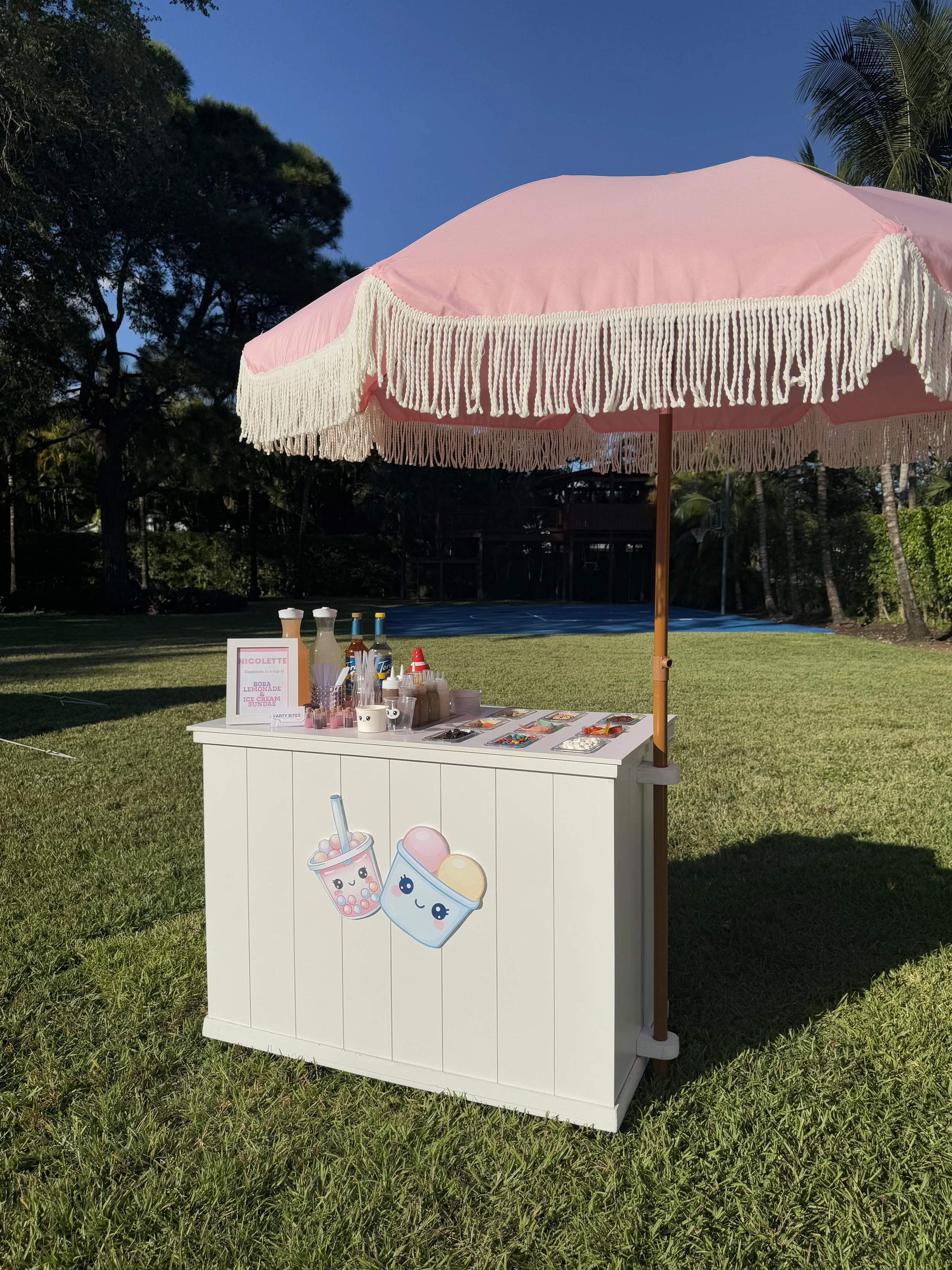 A pink and white lemonade stand with a fringe-trimmed pink umbrella, set up on a grassy field under a clear blue sky, decorated with cute cartoon stickers of bubble tea and ice cream, with bottles, cups, and supplies on top.