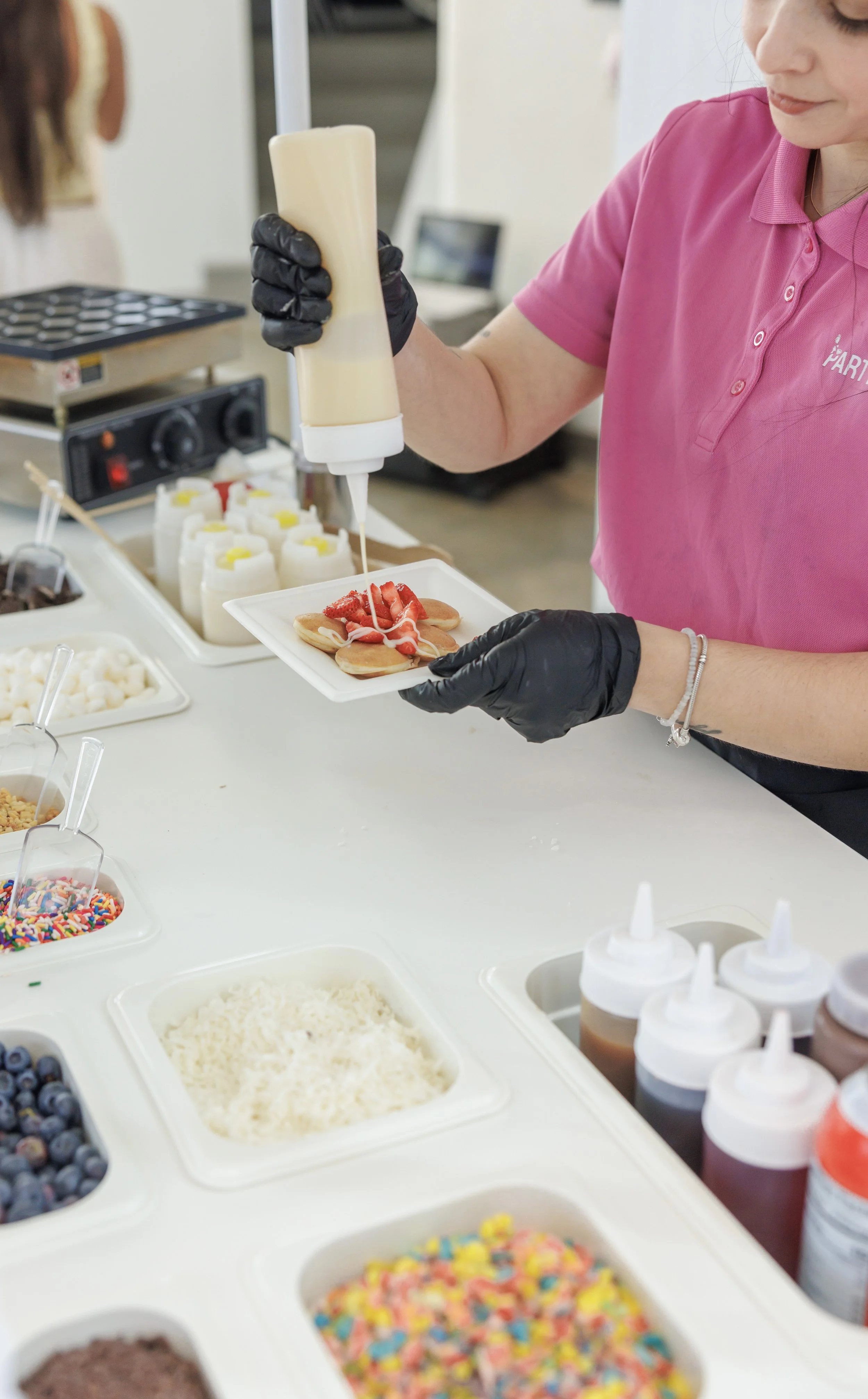 A person in a pink shirt and black gloves decorator making a cookie with strawberries and whipped cream, with various toppings and sauces visible on the counter.