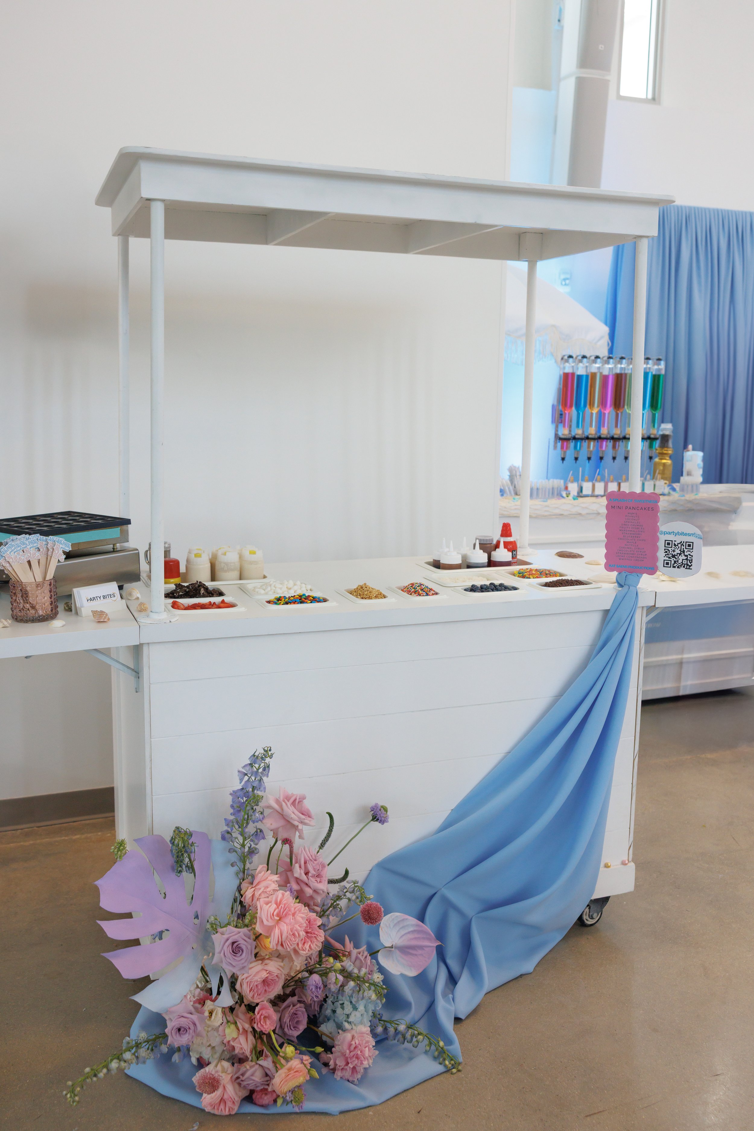Empty white dessert and topping station with colorful sprinkles, syrups, and toppings, decorated with pink and purple flowers with blue drape.