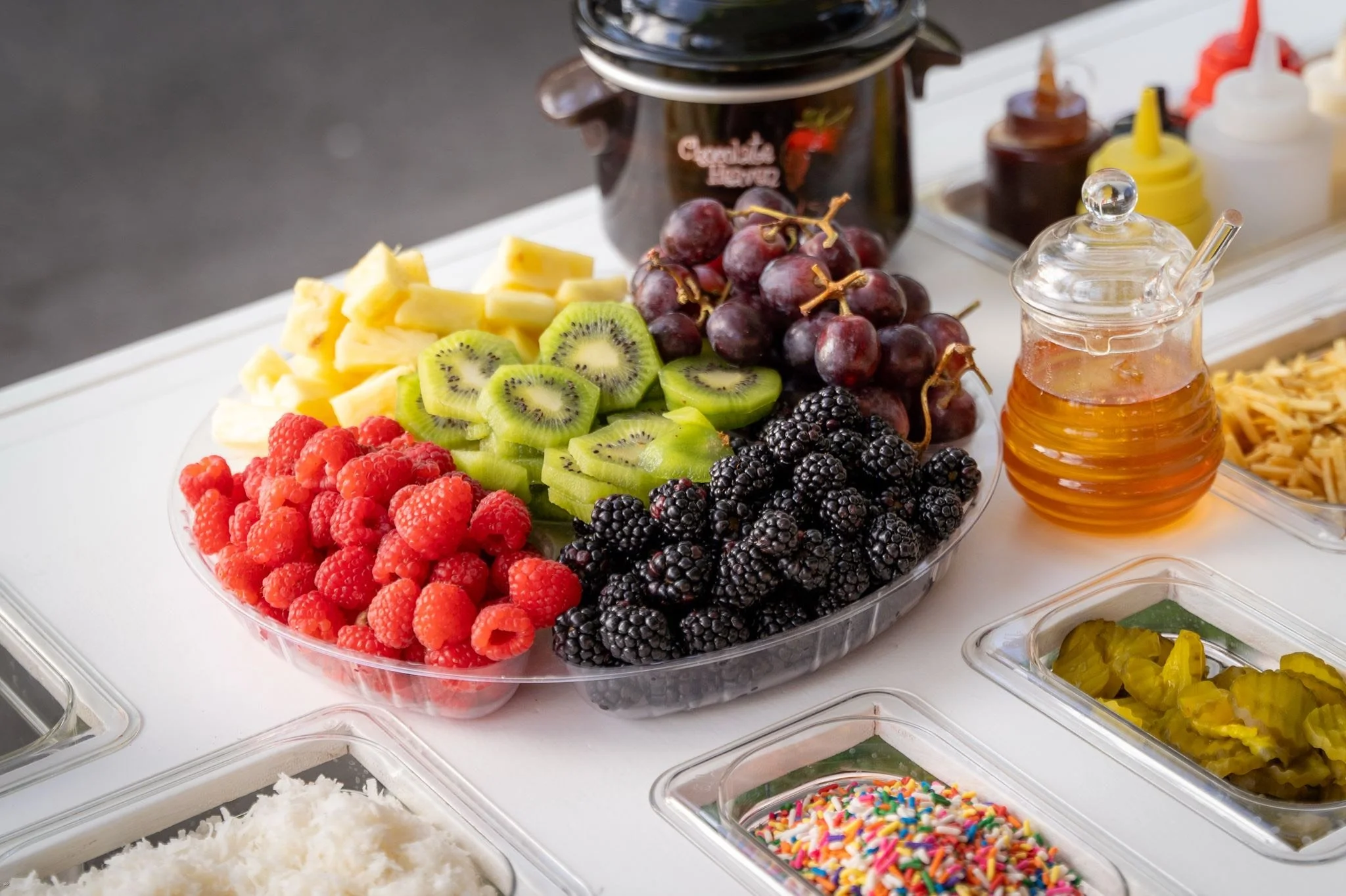 Fresh cut fruit including raspberries, kiwi, pineapple, blackberries, and red grapes on a serving tray at a buffet table.