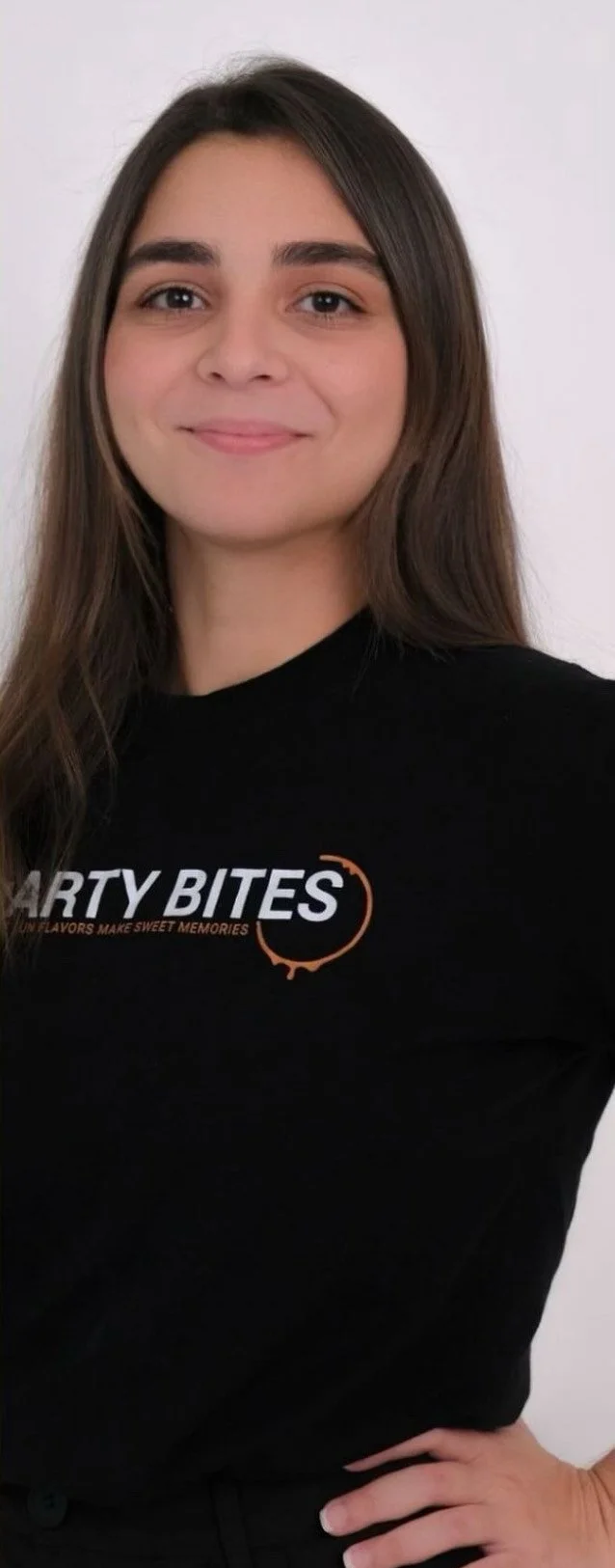 A young woman with long brown hair smiling, wearing a black shirt with the words "PARTY BITES" and a tagline underneath, standing against a plain white background.