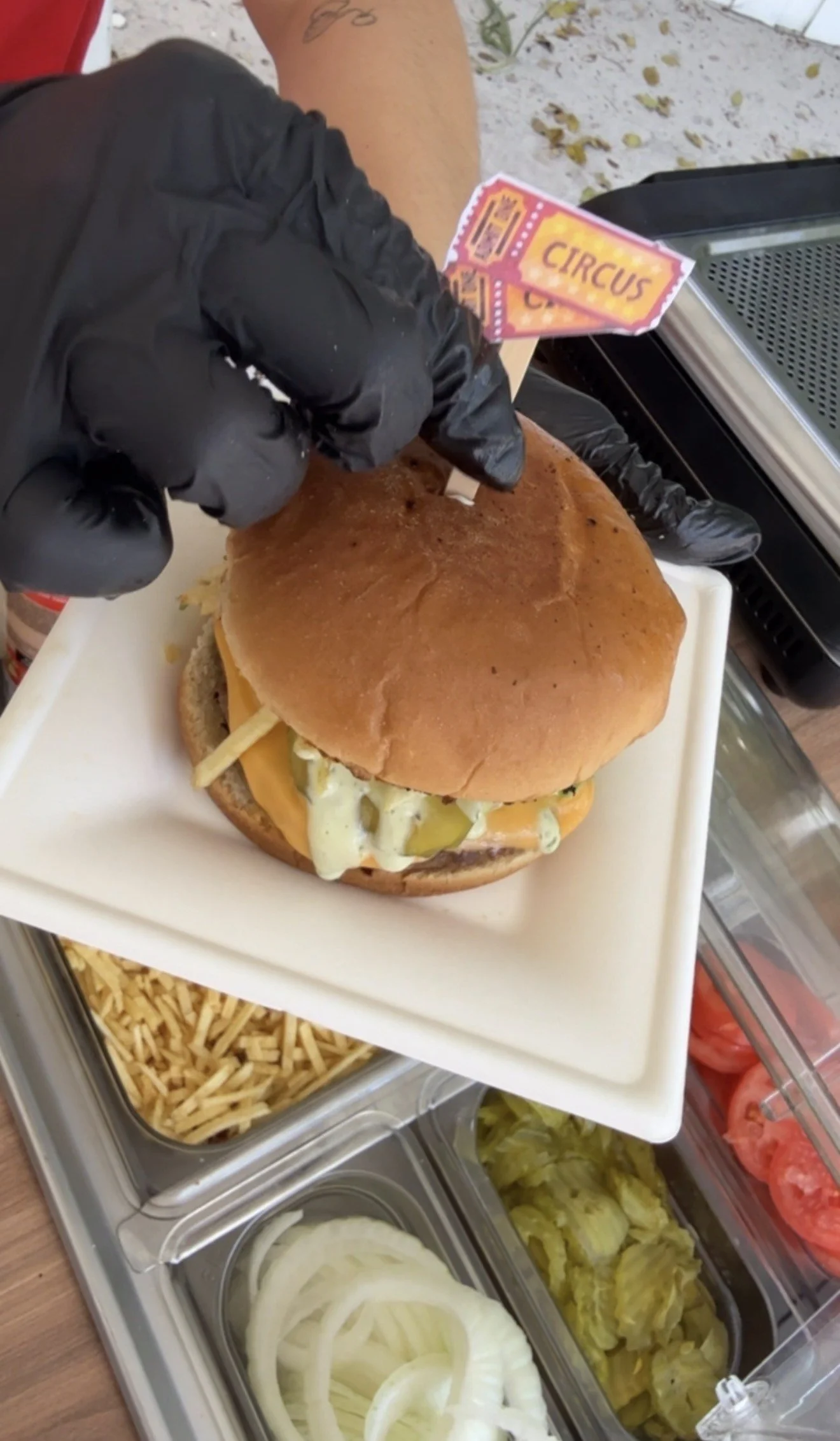A person wearing black gloves is preparing a large sandwich or burger with a bun, cheese, and condiments. The sandwich is on a white tray, and there are containers of sliced onions, pickles, lettuce, and sliced tomatoes nearby.