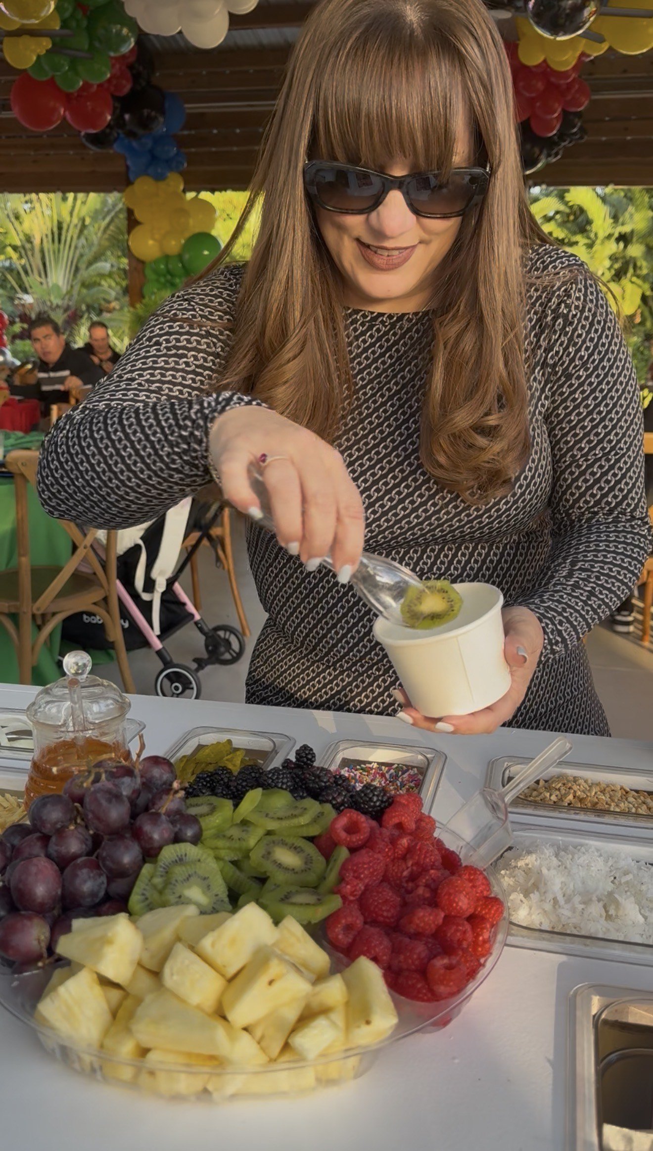 Woman wearing sunglasses serving ice cream with kiwi and other toppings at a fruit and dessert table decorated with balloons.