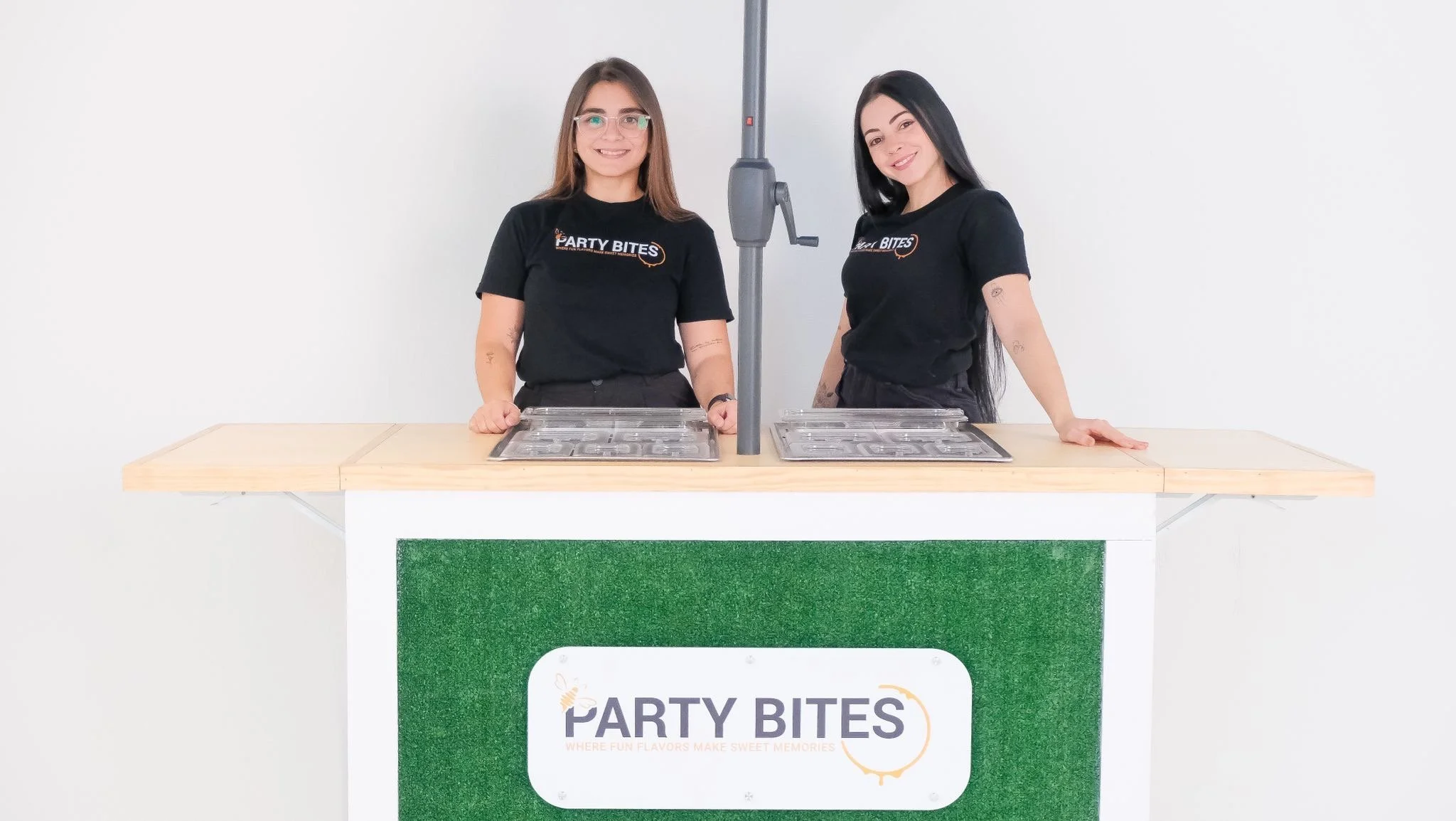 Two women standing behind a party ice cream bar with a green and white counter, both wearing black t-shirts with the 'Party Bites' logo, smiling, in front of a plain white background.