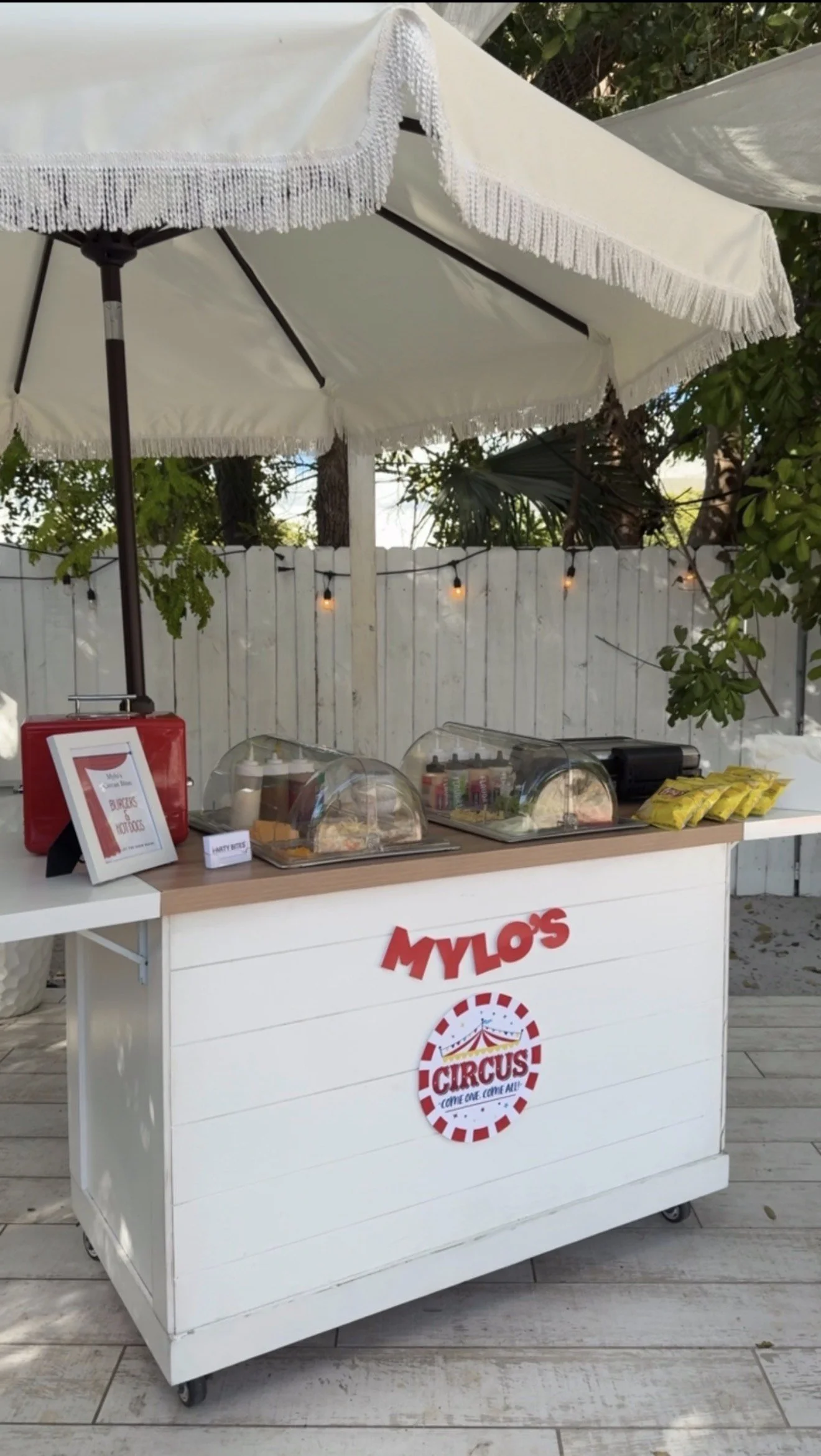 A portable food stand labeled MYLO'S CIRCUS with a white wooden front panel, under a large white umbrella, set up outdoors on a wooden deck, with food containers and snacks on top, surrounded by greenery and string lights.