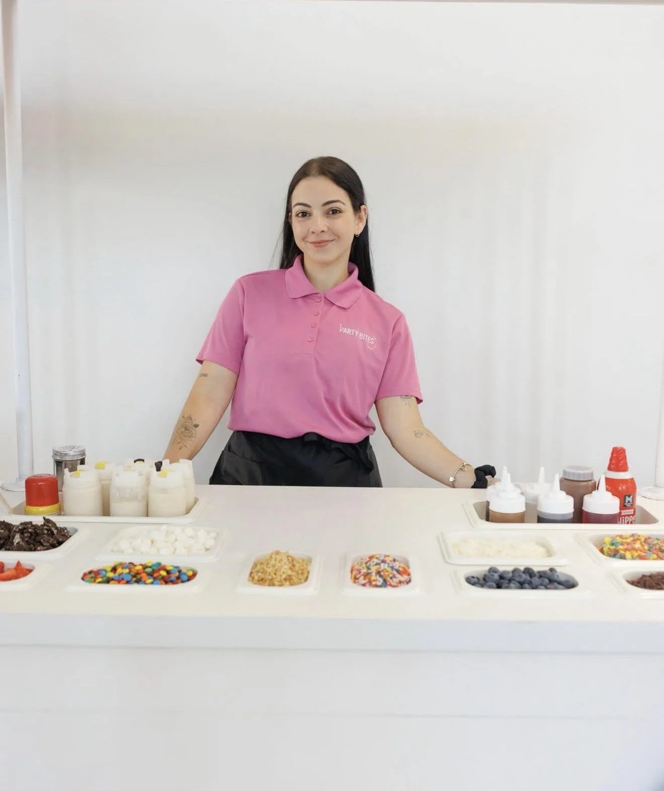 Woman standing behind a table with various toppings and syrups, wearing a pink shirt with the logo 'FARTY BITES'.