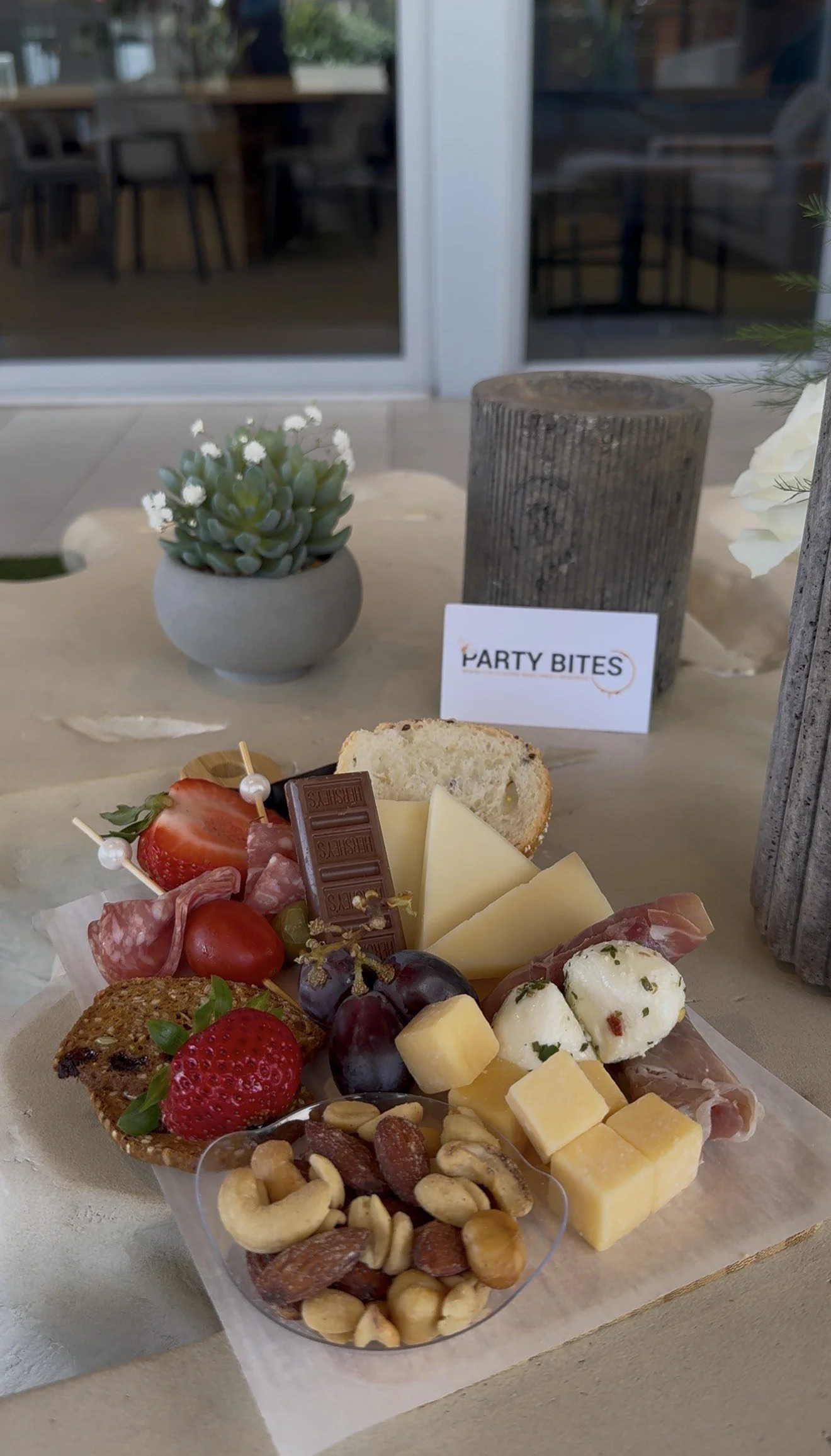 A cheese and charcuterie platter with grapes, strawberries, nuts, sliced bread, and assorted cheeses, placed on a table with a potted succulent plant and a sign that says 'PartY Bites' in the background.