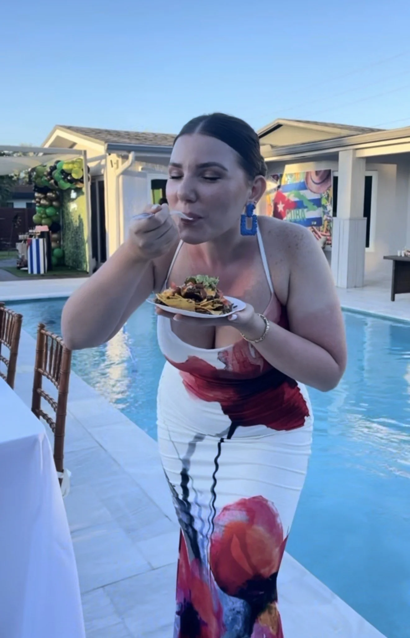 Woman in a floral dress standing by a pool, eating food from a plate during an outdoor gathering, with party decorations in the background.