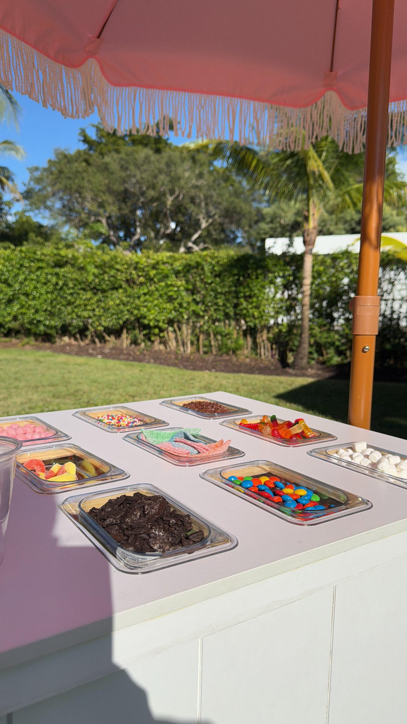 A variety of candies and chocolates in small containers on a white table under pink umbrella in outdoor garden setting.