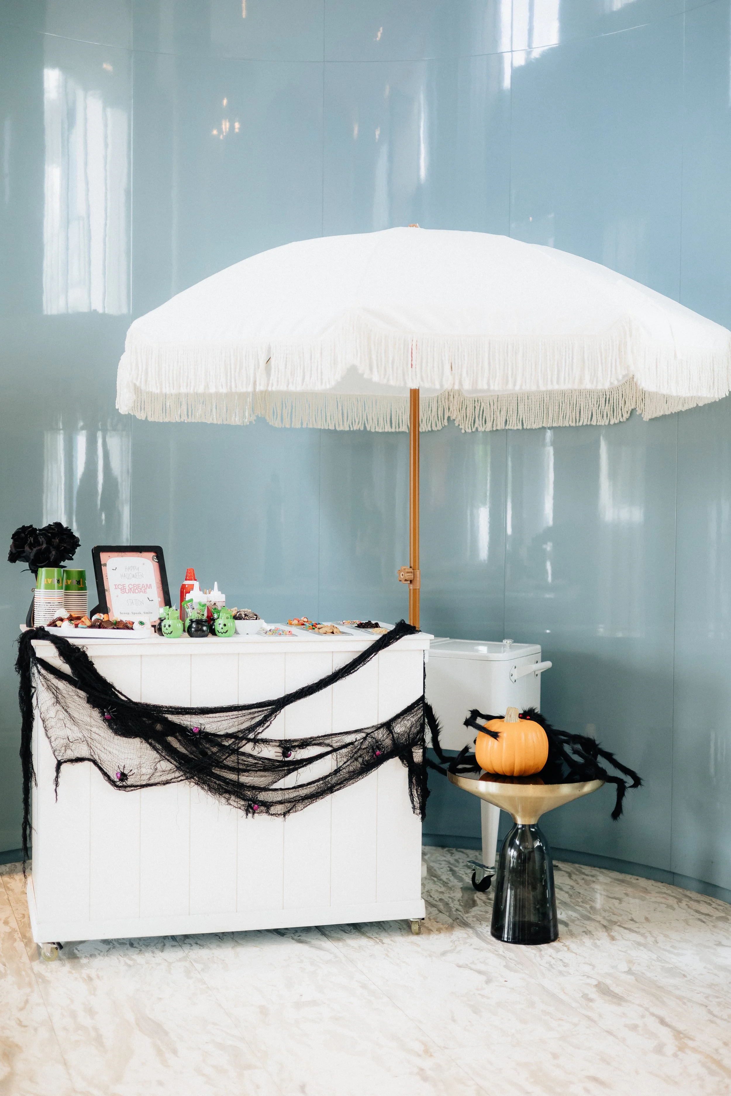 A Halloween-themed ice cream sundae stand decorated with black spider web garlands and pumpkins, shaded by a large white umbrella.
