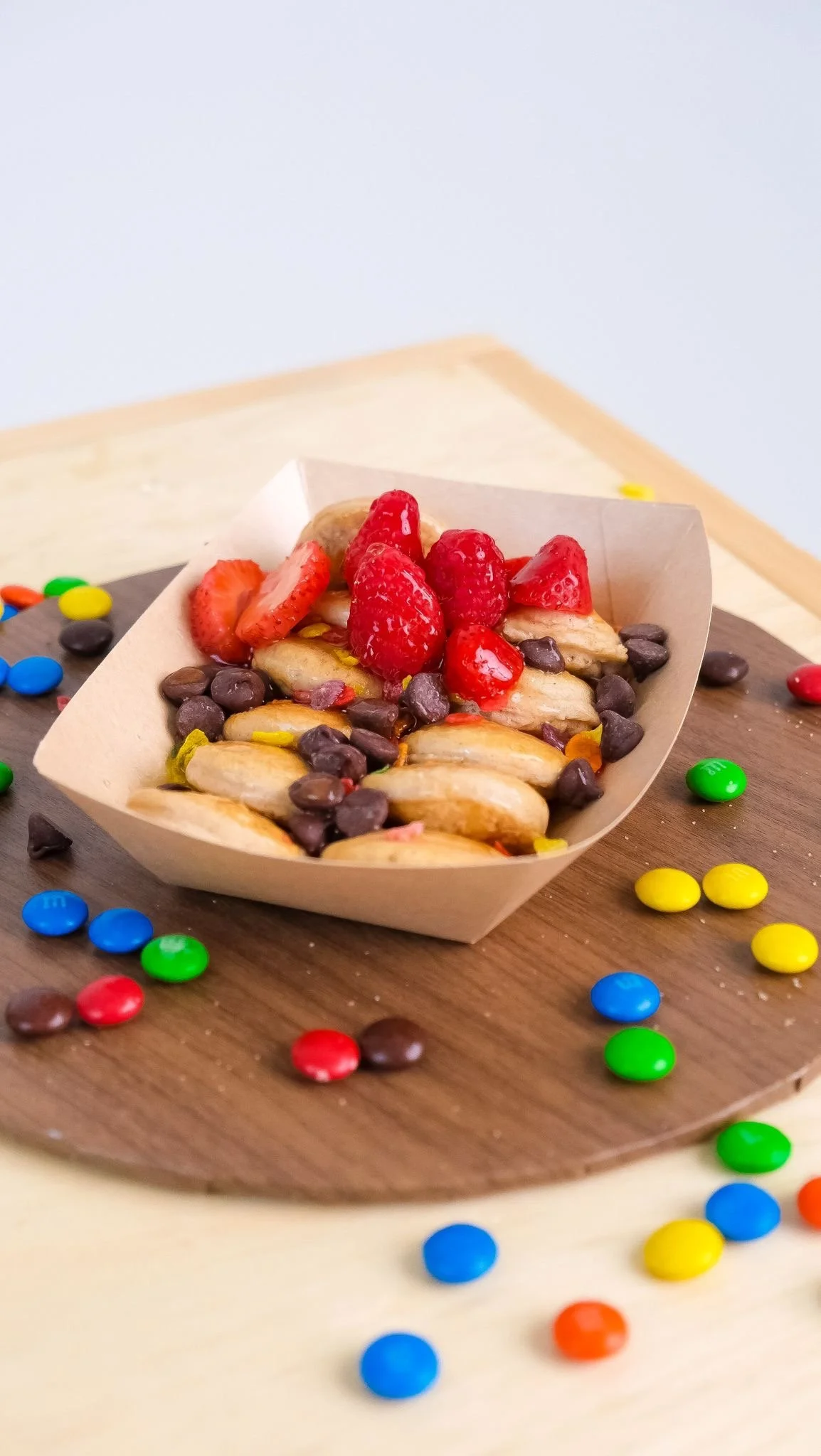 A small paper tray filled with strawberries, bananas, chocolate chips, and colorful candy-coated chocolates on a wooden table with scattered jelly beans.