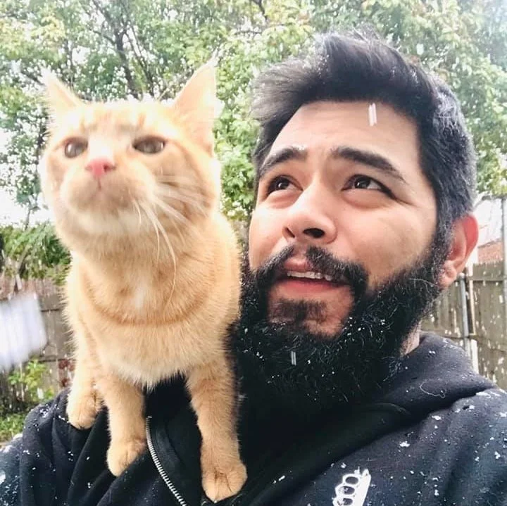 A man with a beard and a black hoodie taking a selfie outdoors with a ginger cat perched on his shoulder, with green trees and a wooden fence in the background.