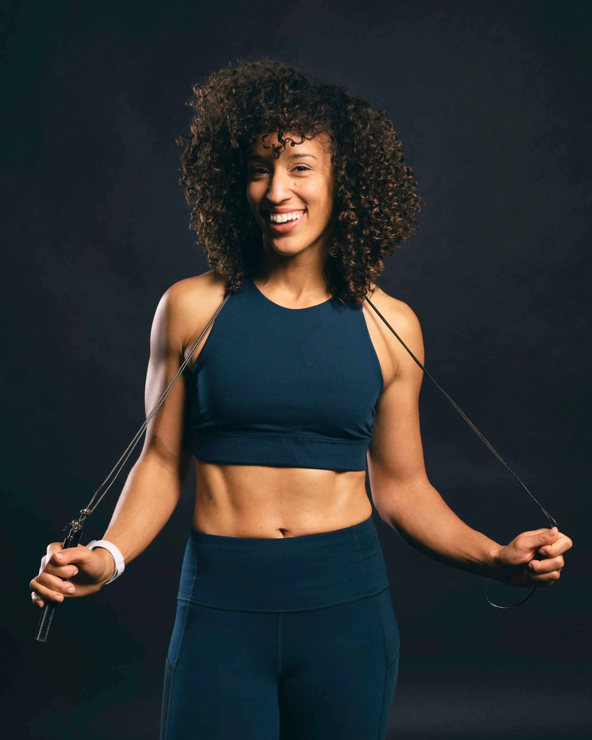 A woman with curly hair smiling while holding a jump rope, wearing a dark blue athletic outfit.
