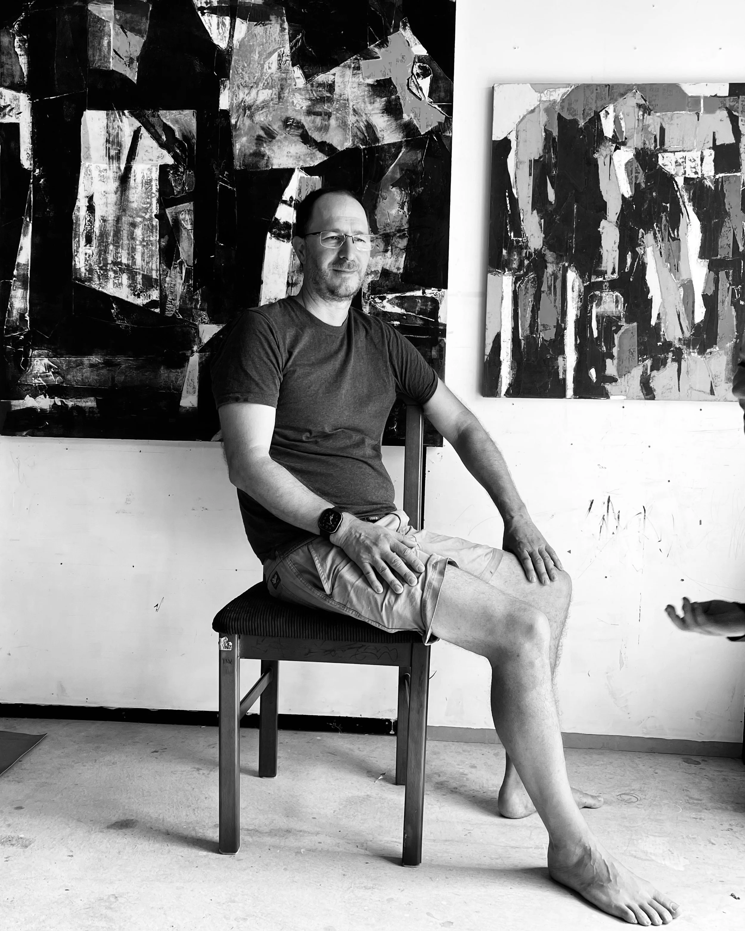 A man sitting on a chair indoors in front of abstract paintings, wearing a black t-shirt and shorts, with one leg extended and the other bent, with his hands resting on his legs.