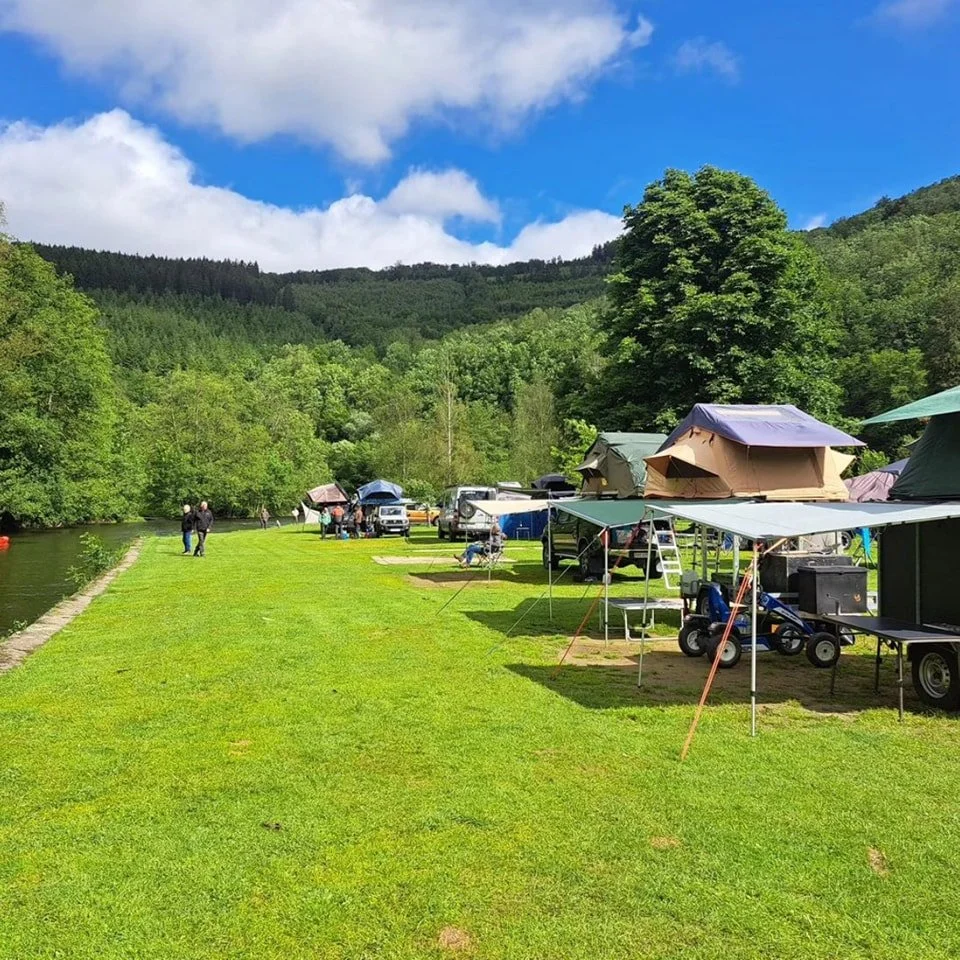 Camping tents set up along a grassy riverside in a lush green forest with mountain hills in the background and a partly cloudy sky.
