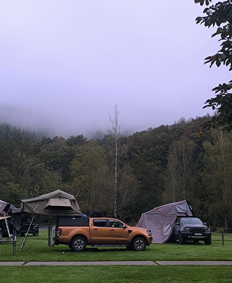 Camping scene with two pickup trucks and tents set up on a grassy field, trees and foggy hills in the background.