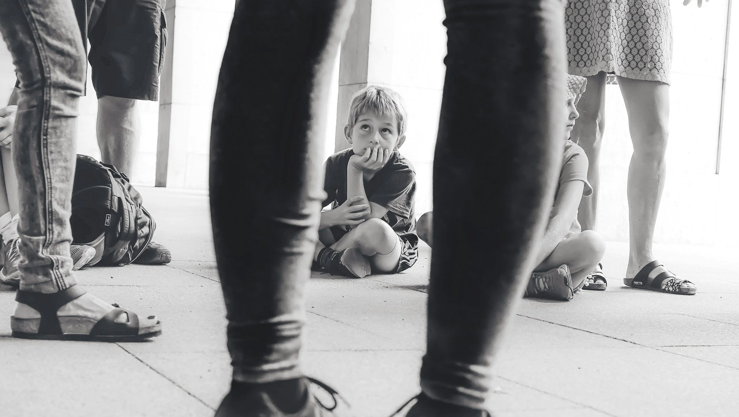 A young boy sitting on the floor, surrounded by adults, resting his chin on his hand and looking upward, with several people standing around him