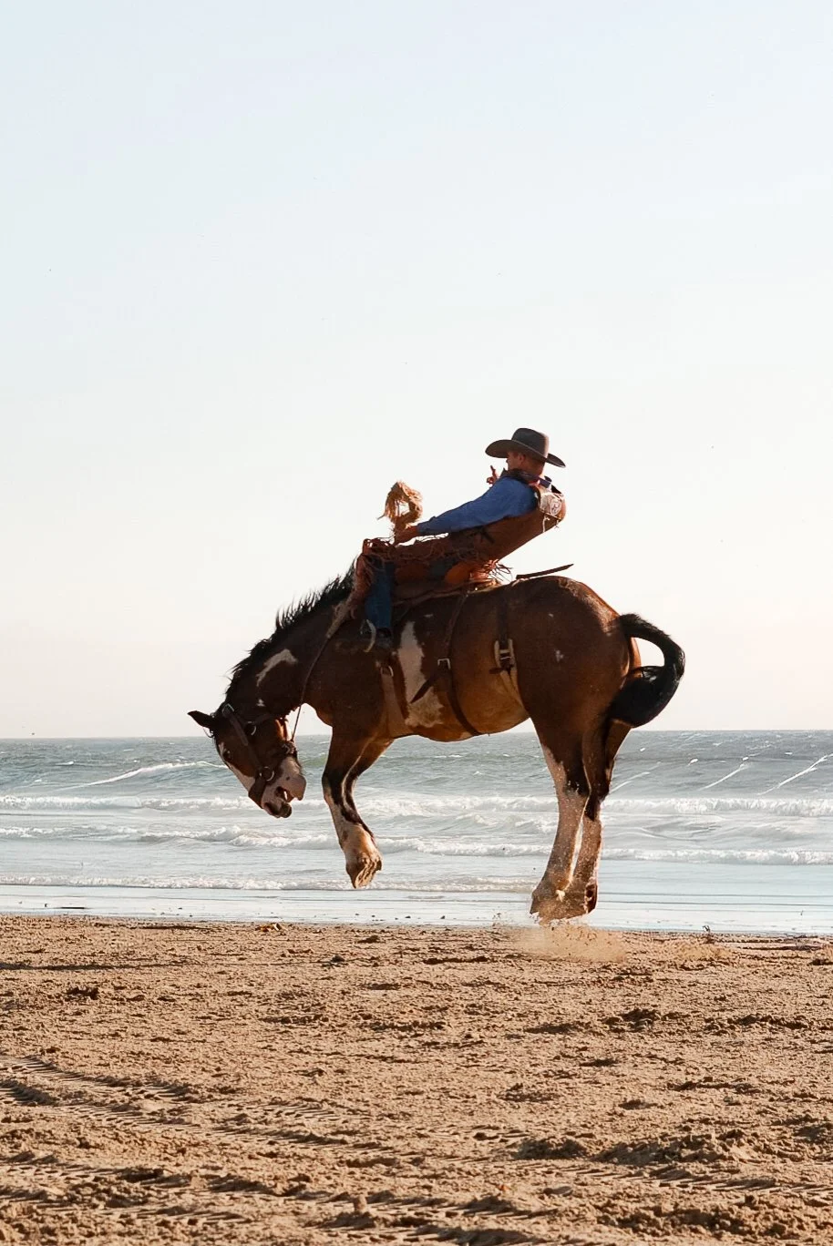 A cowboy riding a bucking horse on a sandy beach near the ocean, with waves in the background.