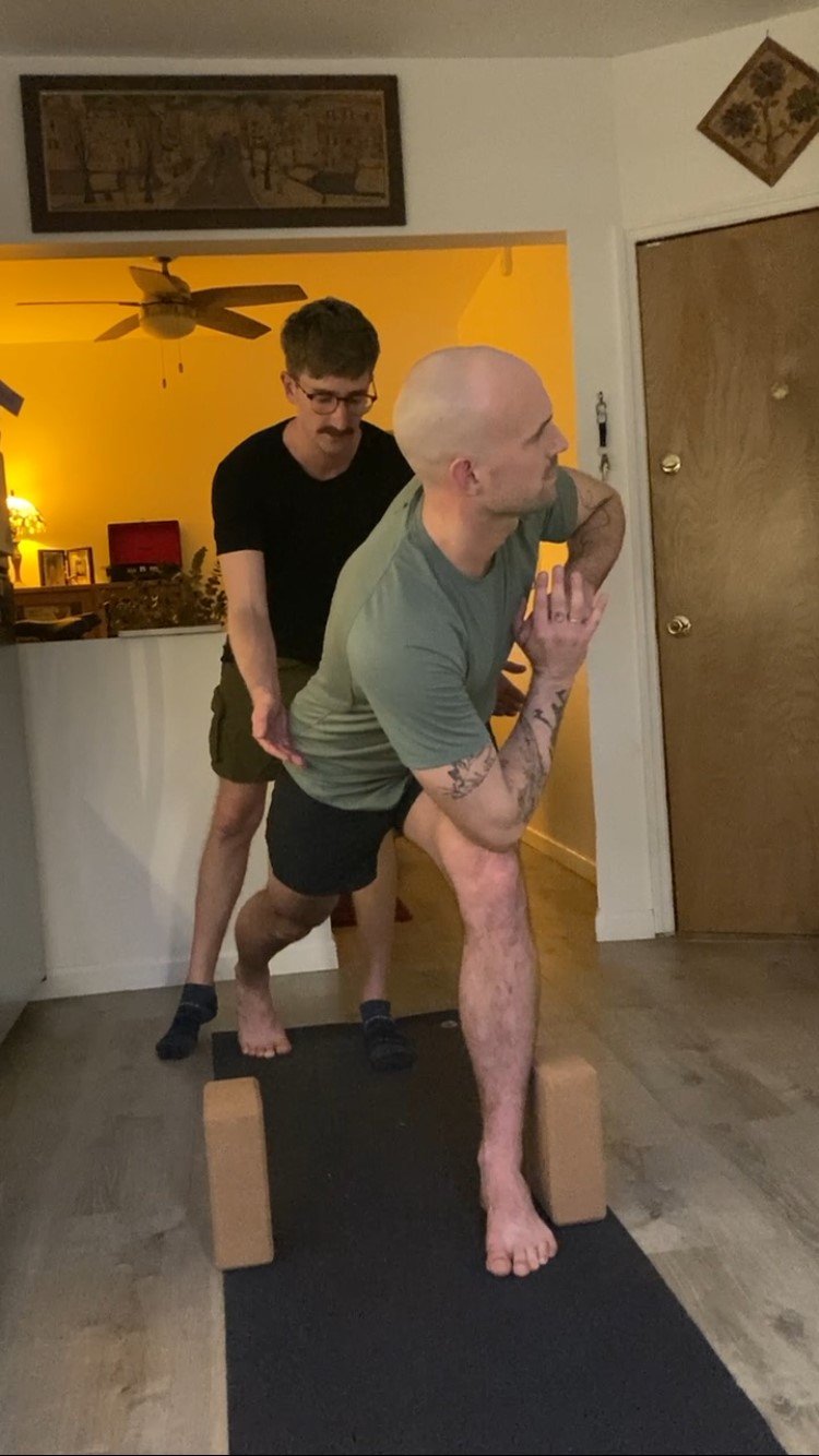 Two men practicing yoga indoors with one man balancing on yoga blocks and the other manual assisting behind him.