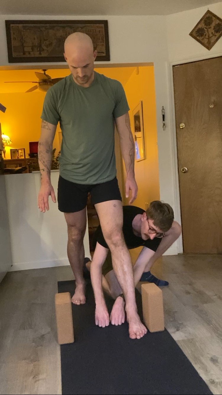 Two men practicing yoga or balancing exercises indoors, with one man standing on a yoga mat on wooden blocks and the other man holding his ankle for support.