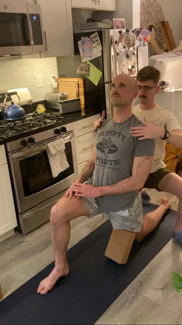 Two men are practicing partner yoga in a kitchen. One man is kneeling on a yoga mat with a cushion under his back, while the other man stands behind him, supporting his shoulder with one hand and touching his chest with the other. They are in a domestic setting with a stove, microwave, refrigerator, and various kitchen items visible.