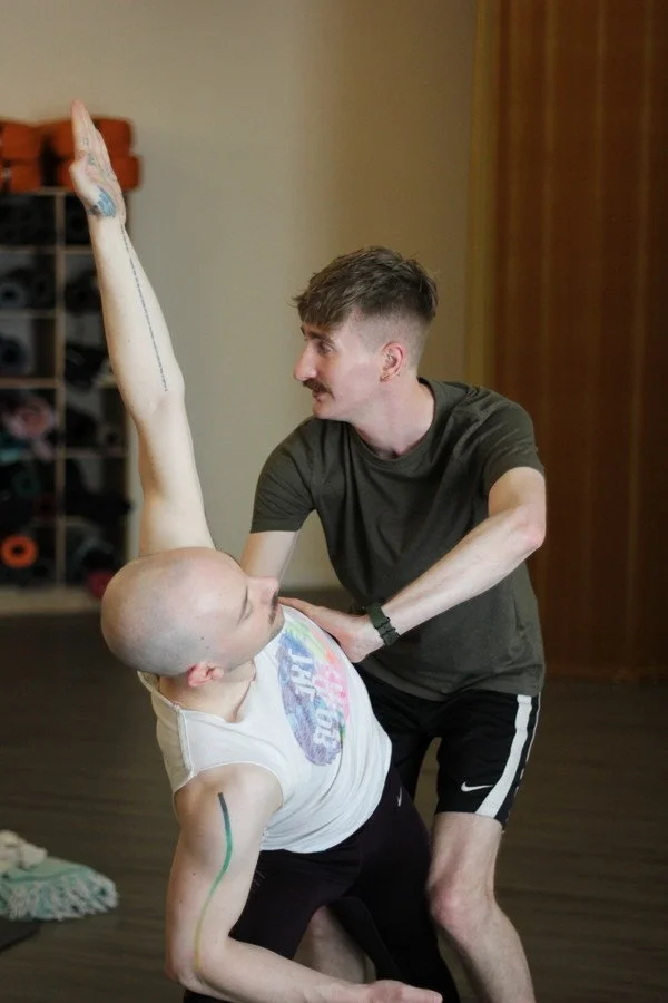 Two men practicing partner yoga or acro yoga in a room, one supporting the other who is extending one arm upwards.