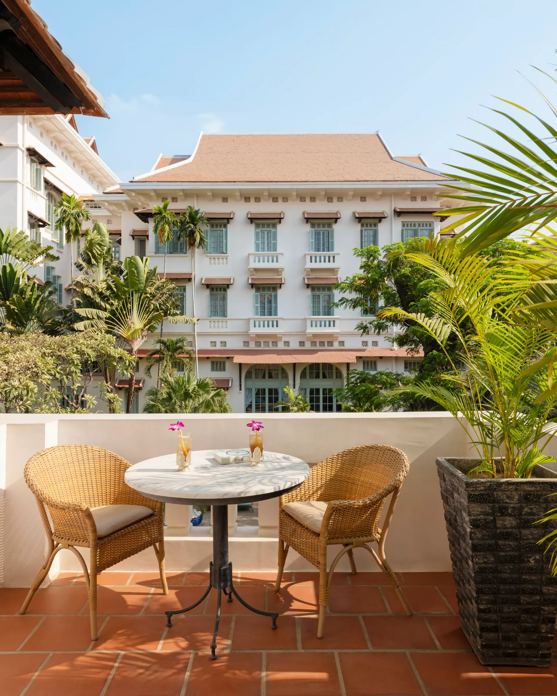 A balcony with a round table, three wicker chairs, and tropical plants in a large planter. Two glasses with drinks and flowers are on the table. A large white building with multiple windows and balconies, and a blue sky in the background.