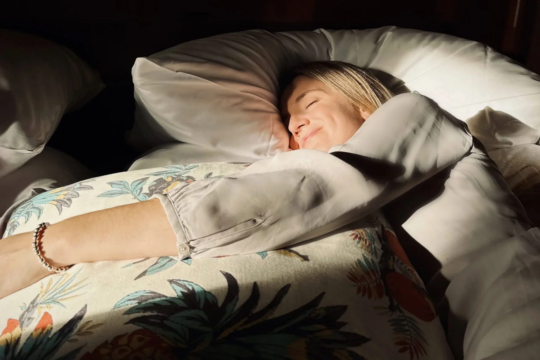 A woman with blond hair lying on a bed, smiling with eyes closed, surrounded by pillows and blankets in a cozy setting.
