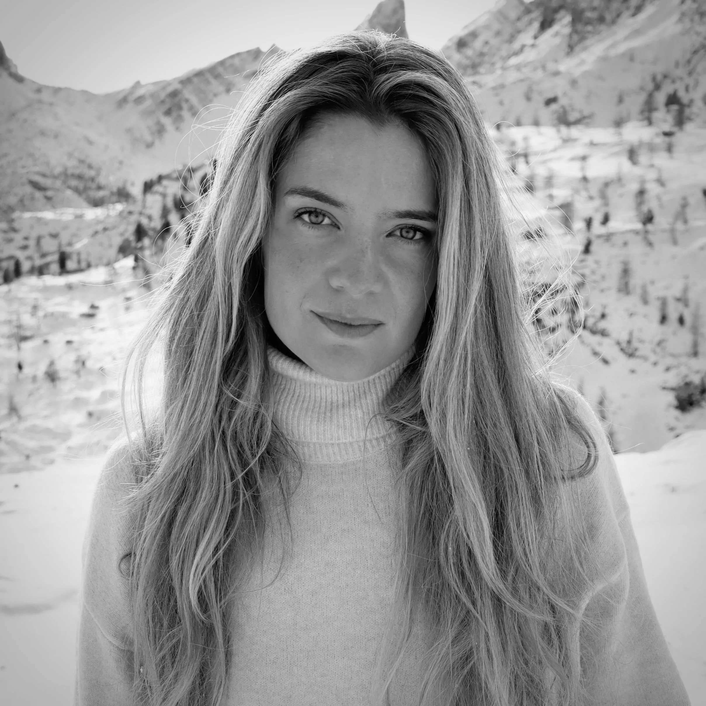 A woman with long, wavy hair and piercing eyes looking at the camera, wearing a turtleneck sweater, snow-covered mountains in the background.