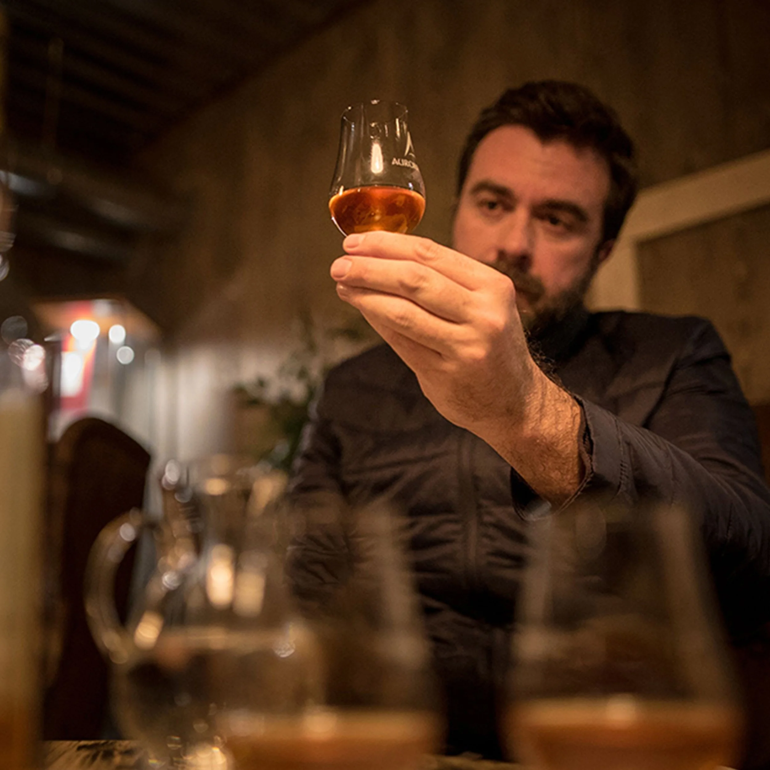 A man in a dark shirt holding a glass of amber-colored beverage, looking at it intently in a dimly lit setting.