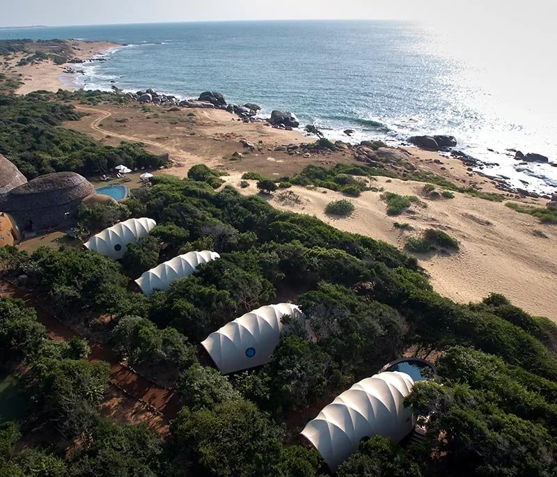Aerial view of eco-friendly domed structures surrounded by greenery near a beach with rock formations and the ocean in the background.