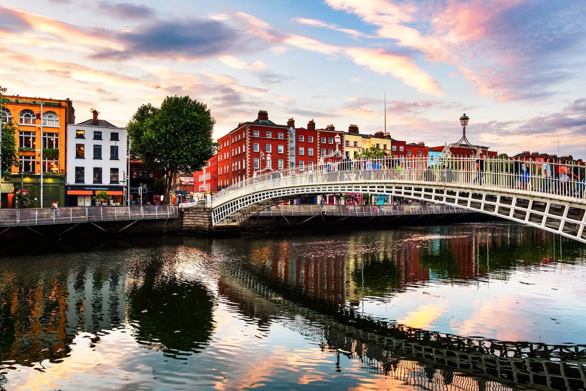 View of a cityscape with colorful buildings, a white arched bridge over water, and a sky with pink and orange clouds at sunset.