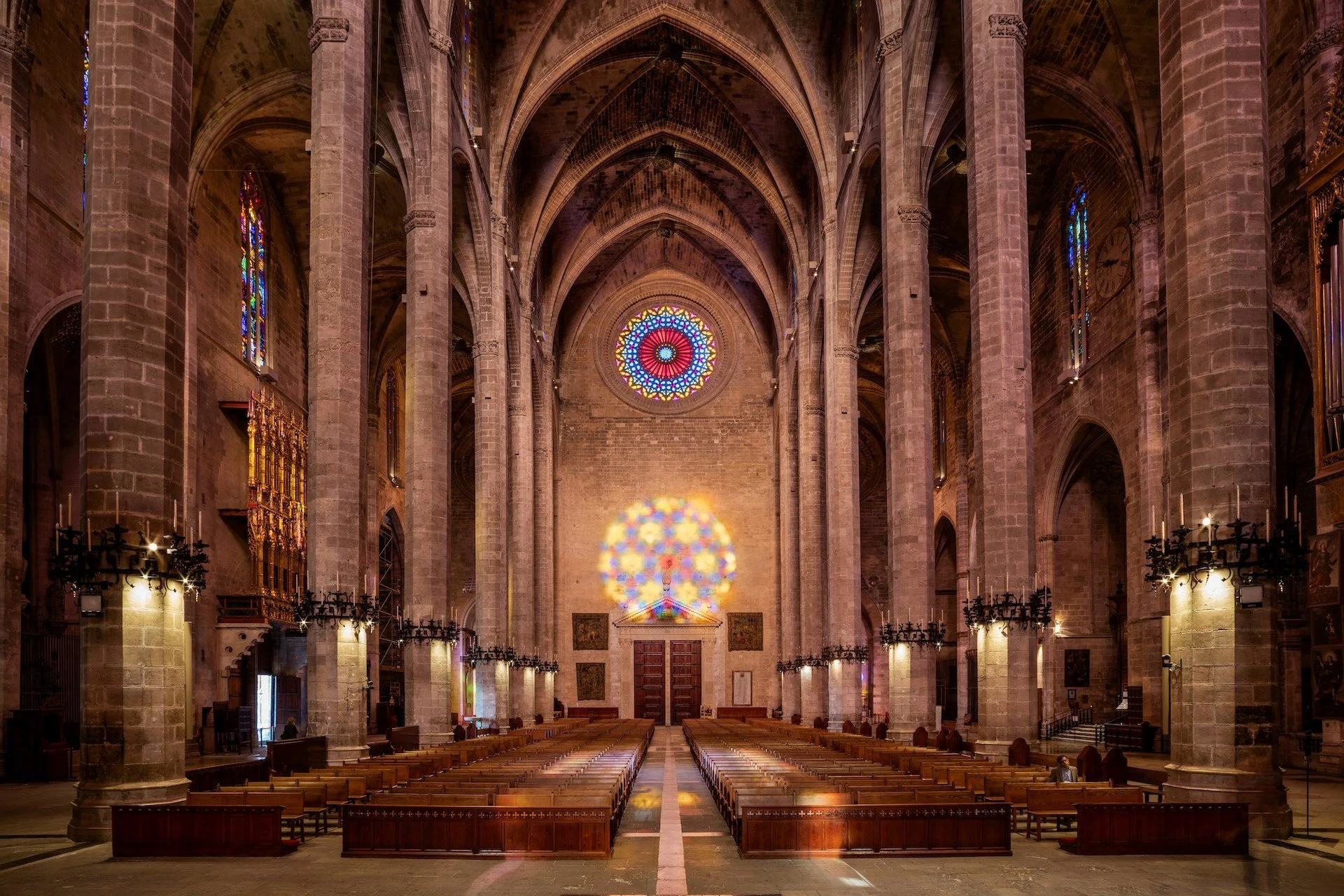 Interior of a Gothic cathedral with high vaulted ceilings, stained glass windows, pillars, and wooden pews, illuminated by natural light and colorful reflections.