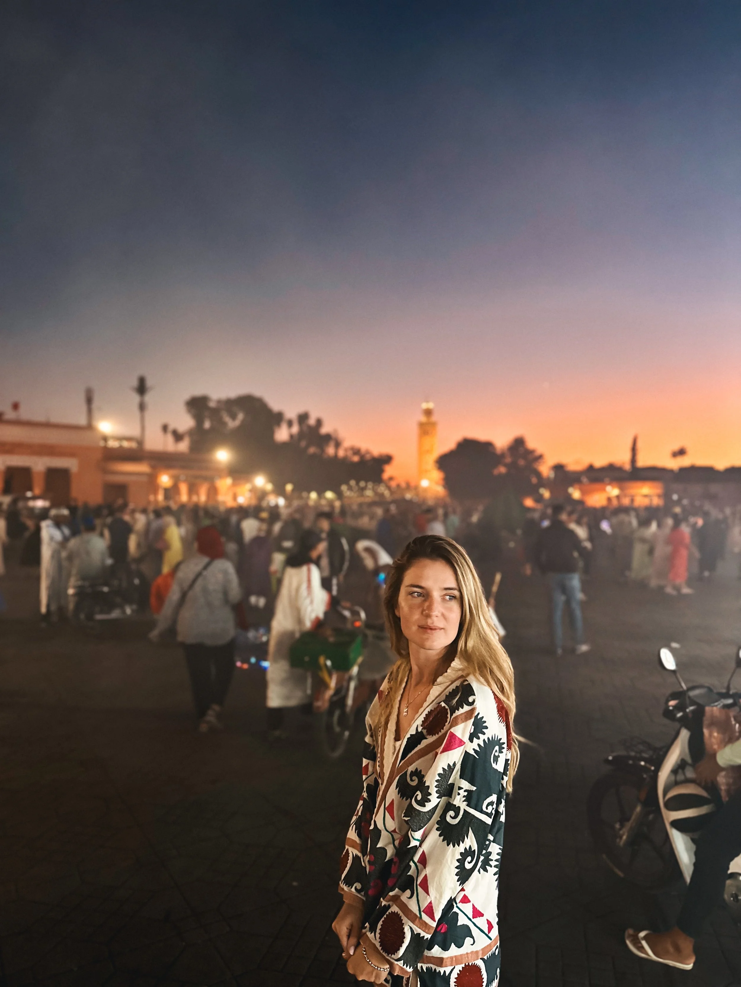 A young woman with long blonde hair standing in a crowded outdoor area at sunset, wearing a colorful patterned jacket, with a cityscape and a tower in the background.