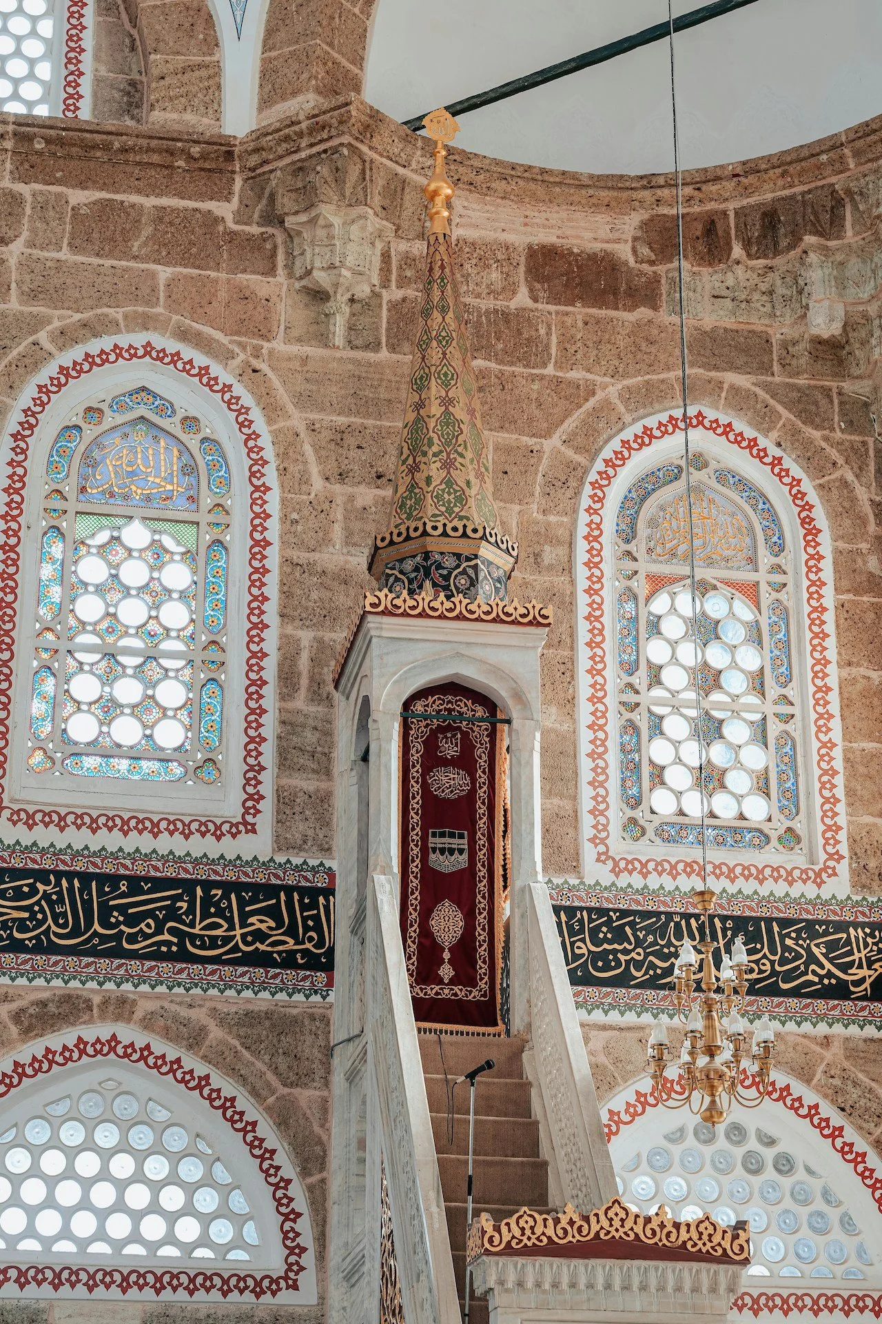 Interior of a mosque with ornate windows, Arabic calligraphy, a pulpit, and a decorative minbar. The architecture features stone walls, detailed glass windows, and a chandelier.