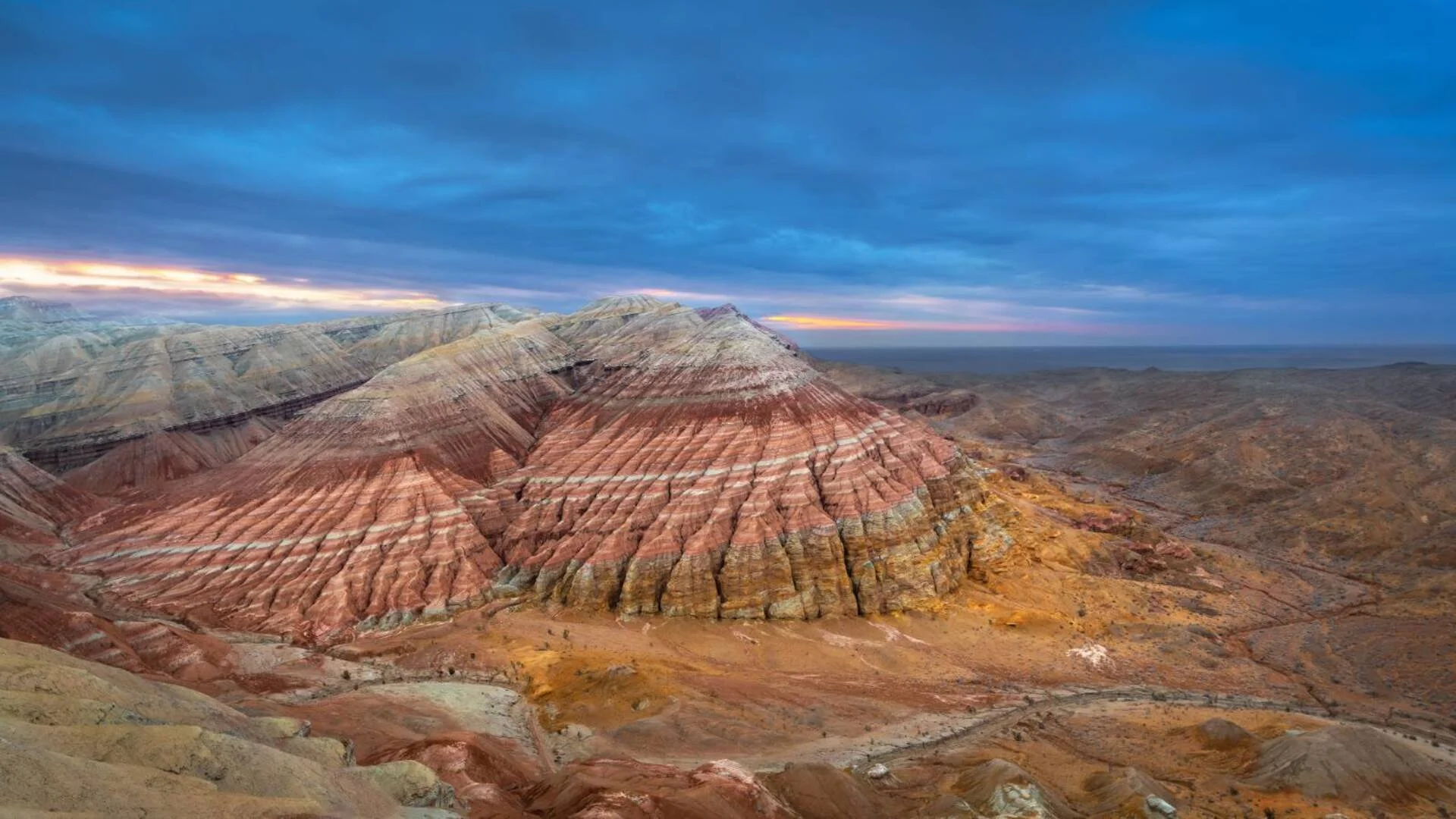 Colorful layered mountains in a desert landscape under a cloudy sky at sunset.