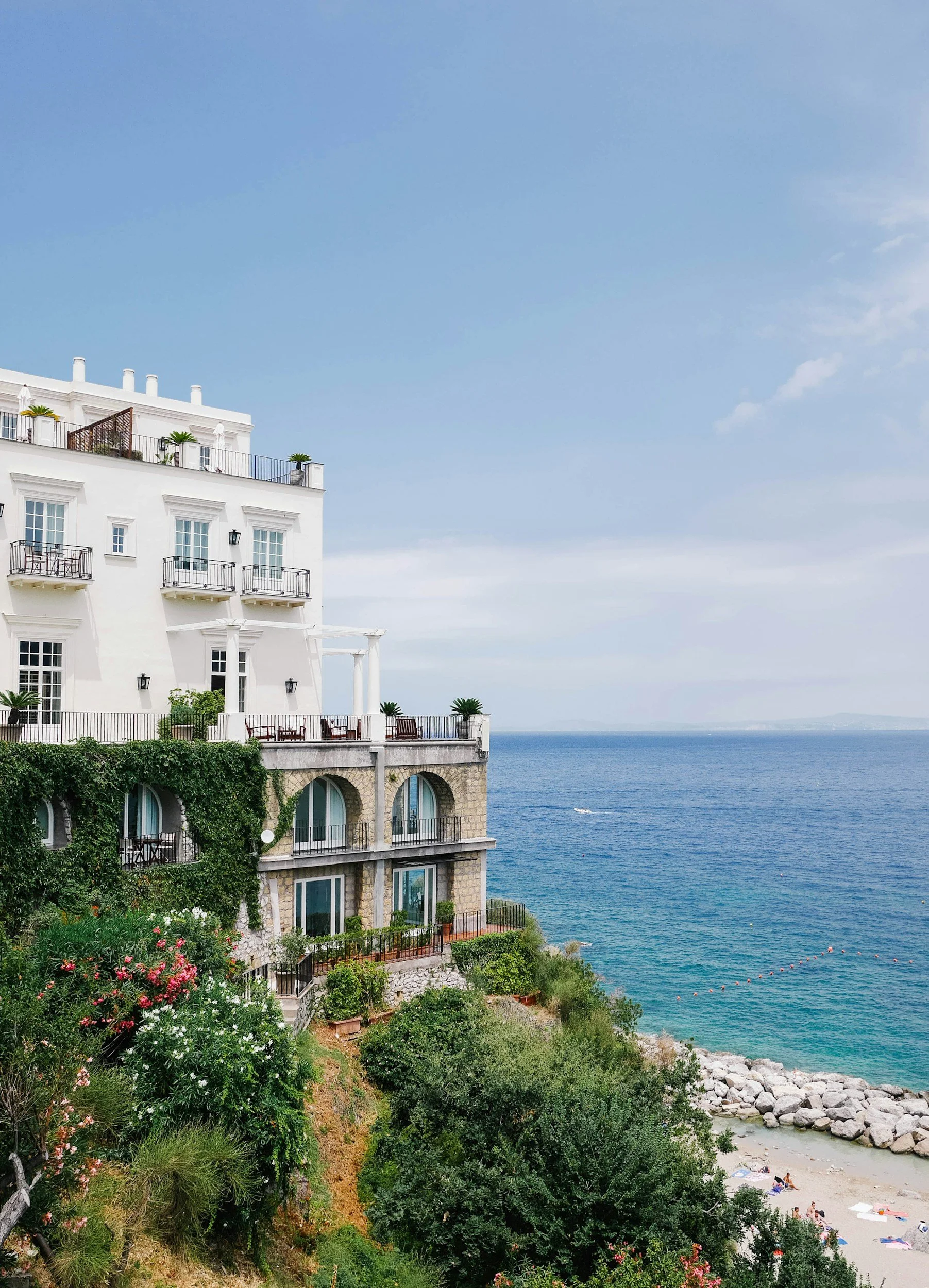 Coastal villa with balconies and lush greenery overlooking a beach and the ocean on a clear day.