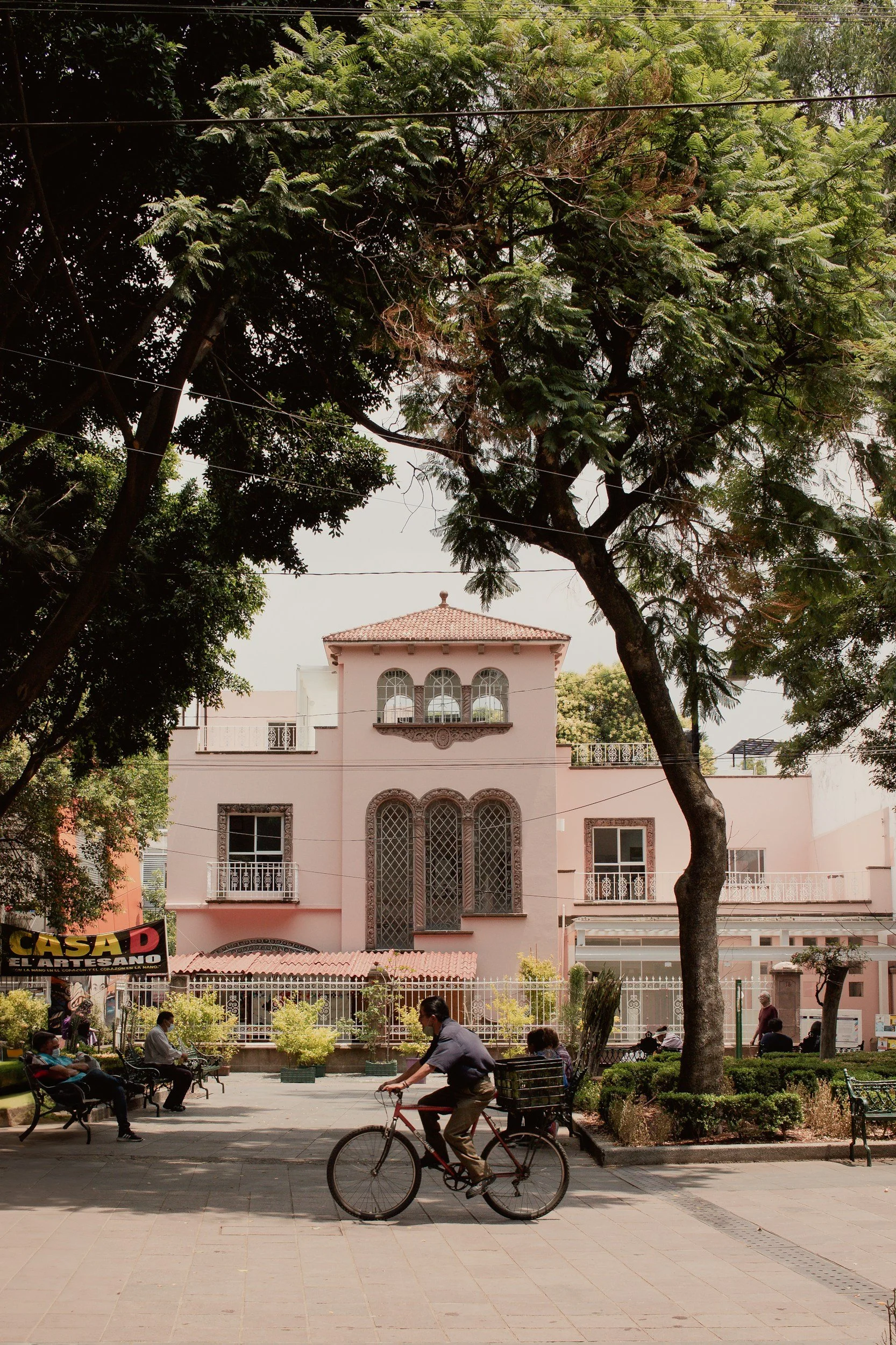 Pink building with ornate windows and balconies surrounded by trees; a cyclist rides past while people sit on benches in a plaza.