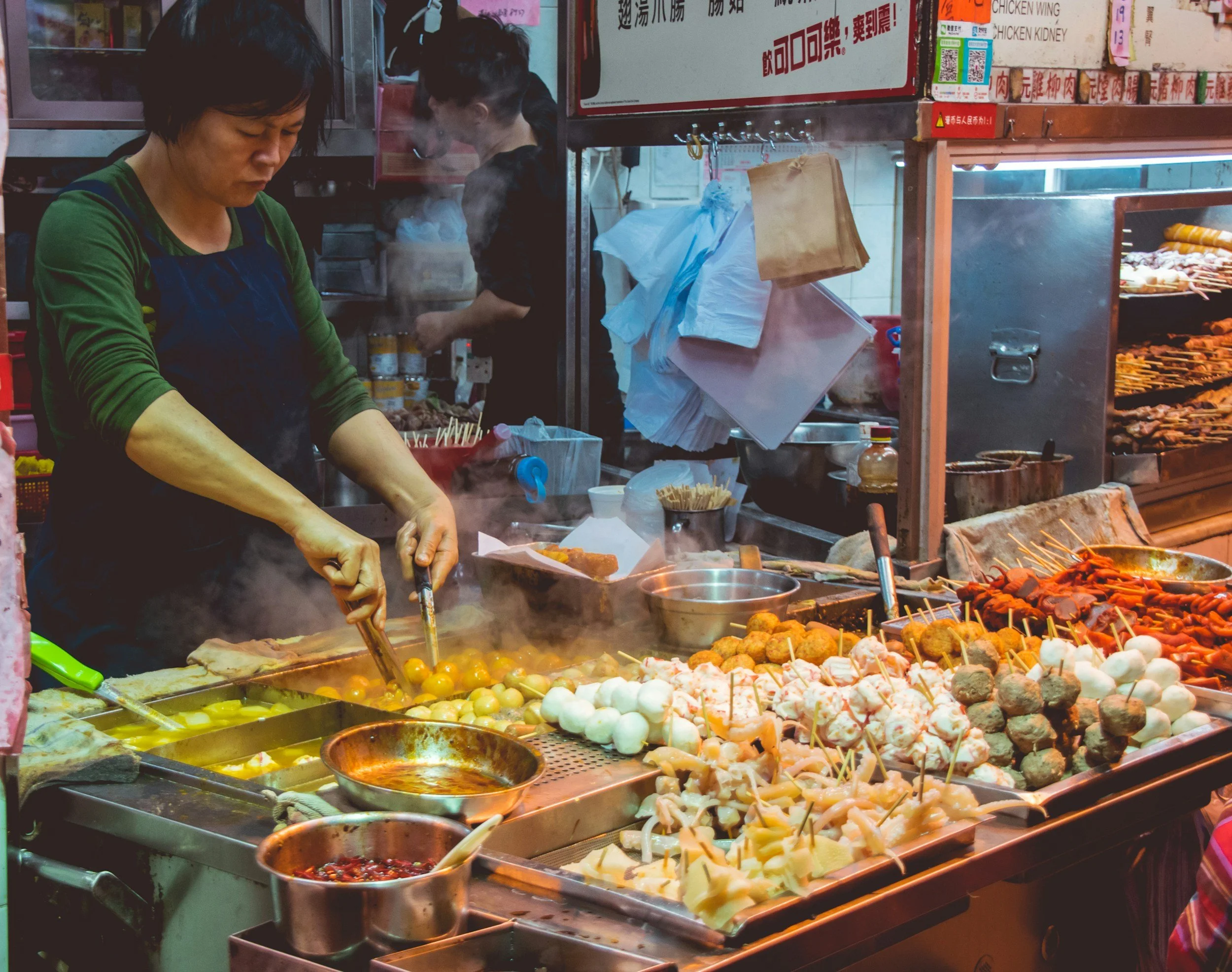 Person cooking street food at a market stall, with various skewers and dishes.