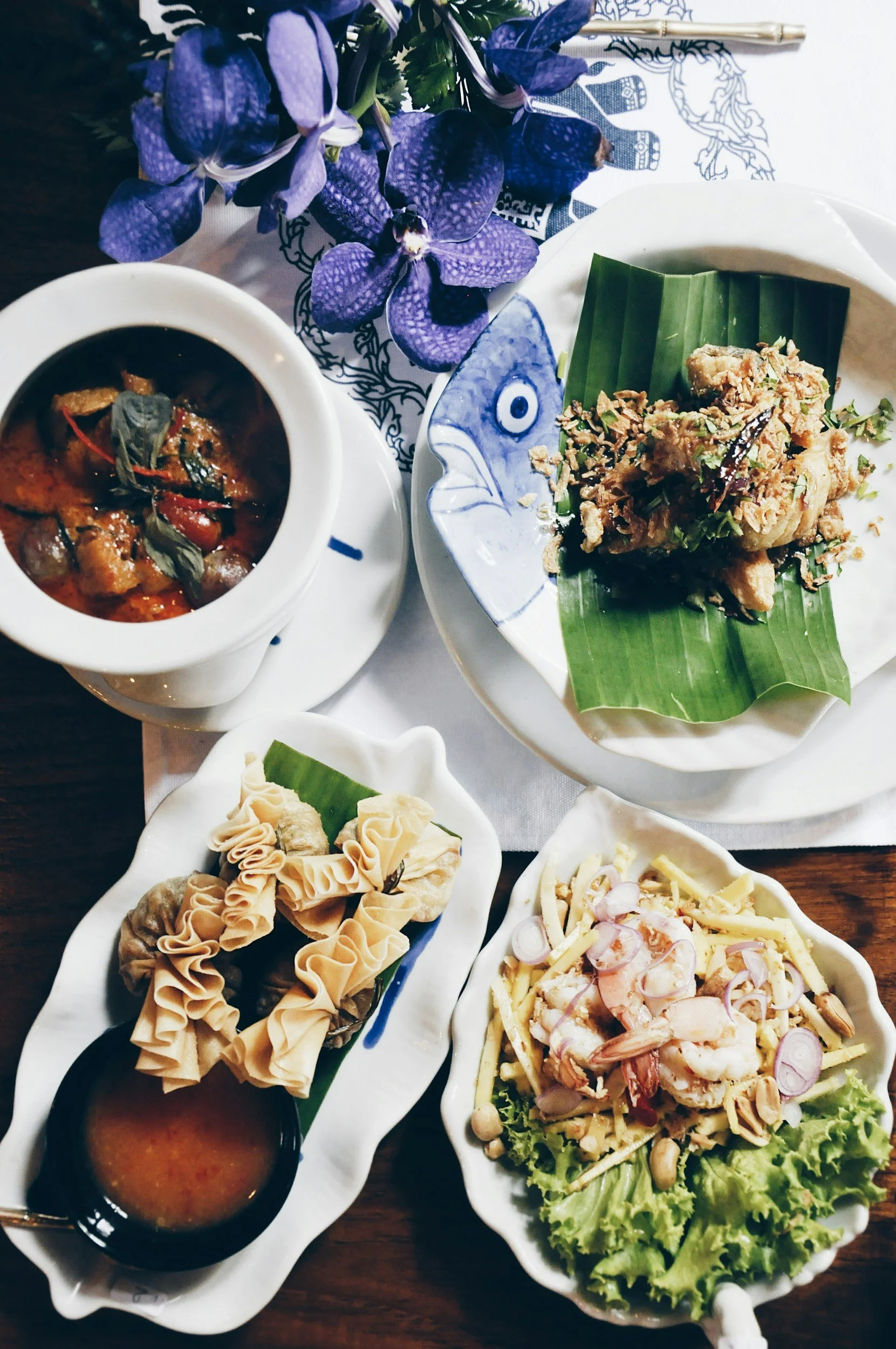 Assorted Thai dishes including a curry in a ceramic bowl, fried dish with crispy onions on banana leaf, dumplings with dipping sauce, and a salad with shrimp and peanuts, accompanied by purple flowers.